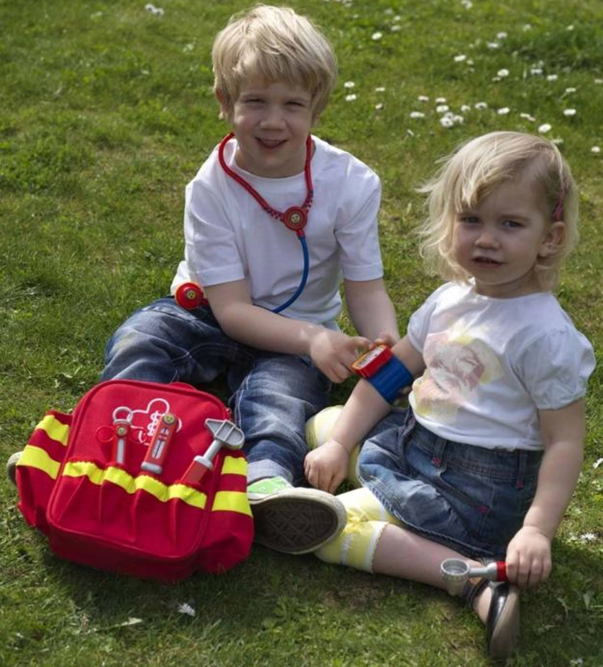 Dwoje dzieci bawiących się w lekarza na trawie, z czerwoną torbą medyczną i zabawkami.