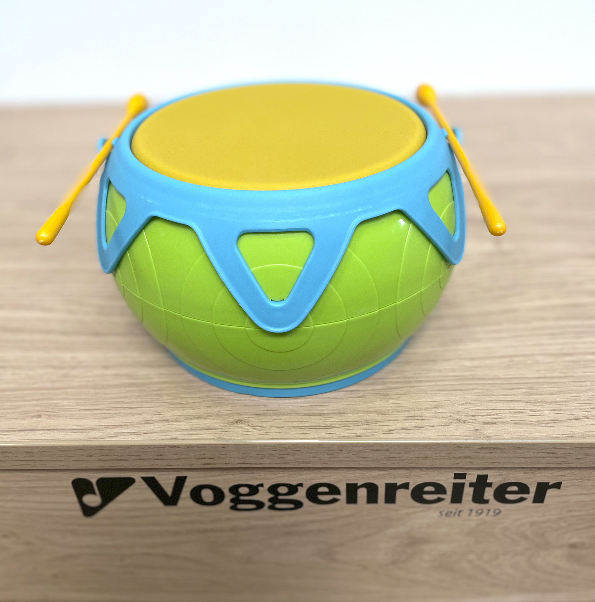 Eine grüne und blaue Spielzeugtrommel mit gelben Trommelstöcken. Sie steht auf einer Holzoberfläche.