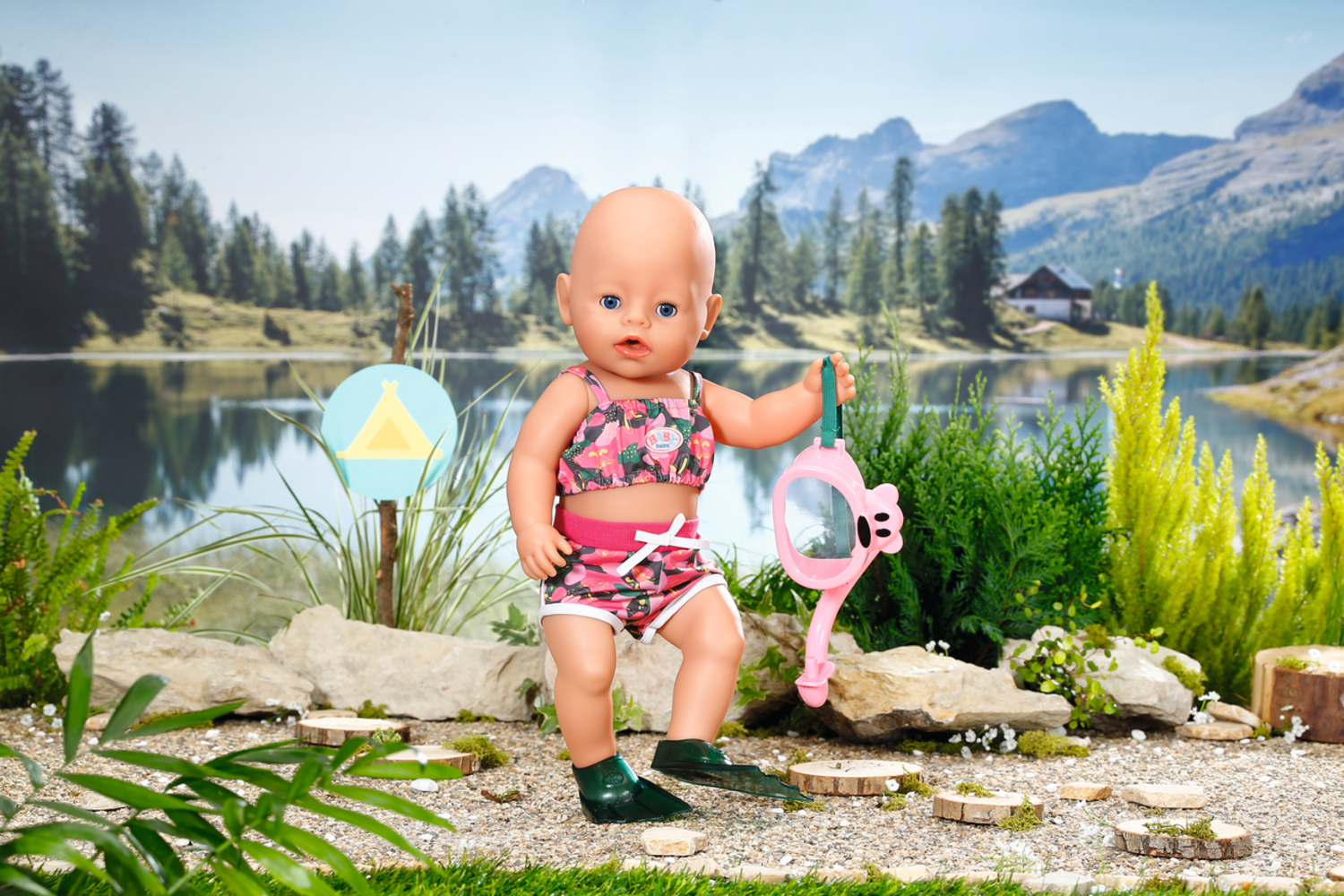 Una muñeca en traje de baño sosteniendo un esnórquel rosa. El fondo es un lago y montañas.