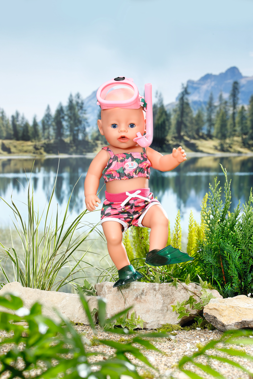 Una muñeca en traje de baño con equipo de snorkel. El fondo es un lago y montañas con un letrero de campamento.