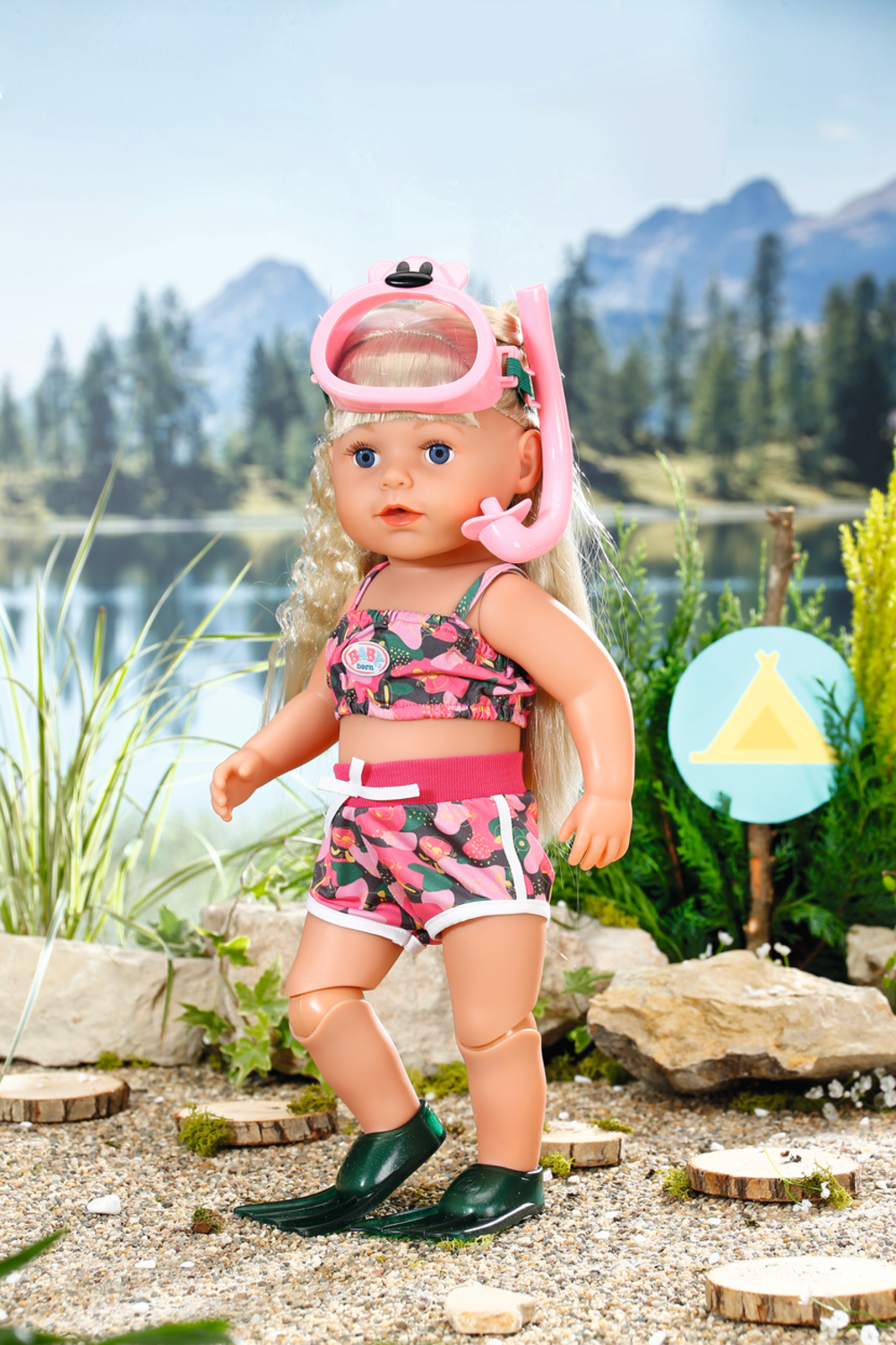 Una muñeca con equipo de snorkel, de pie cerca de un lago con montañas al fondo.