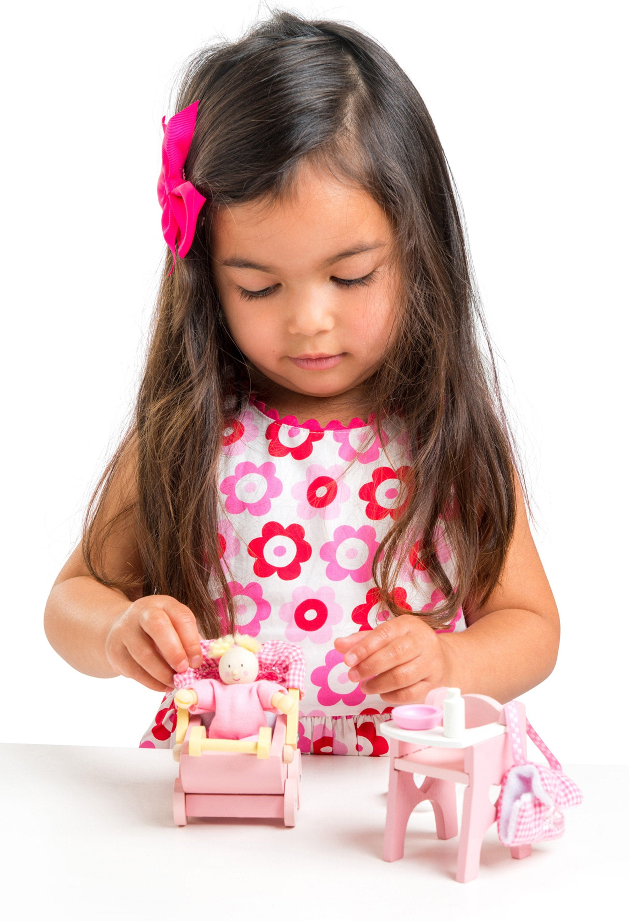 Młoda dziewczyna z długimi brązowymi włosami i różową kokardą bawi się lalką i mebelkami dla lalek.