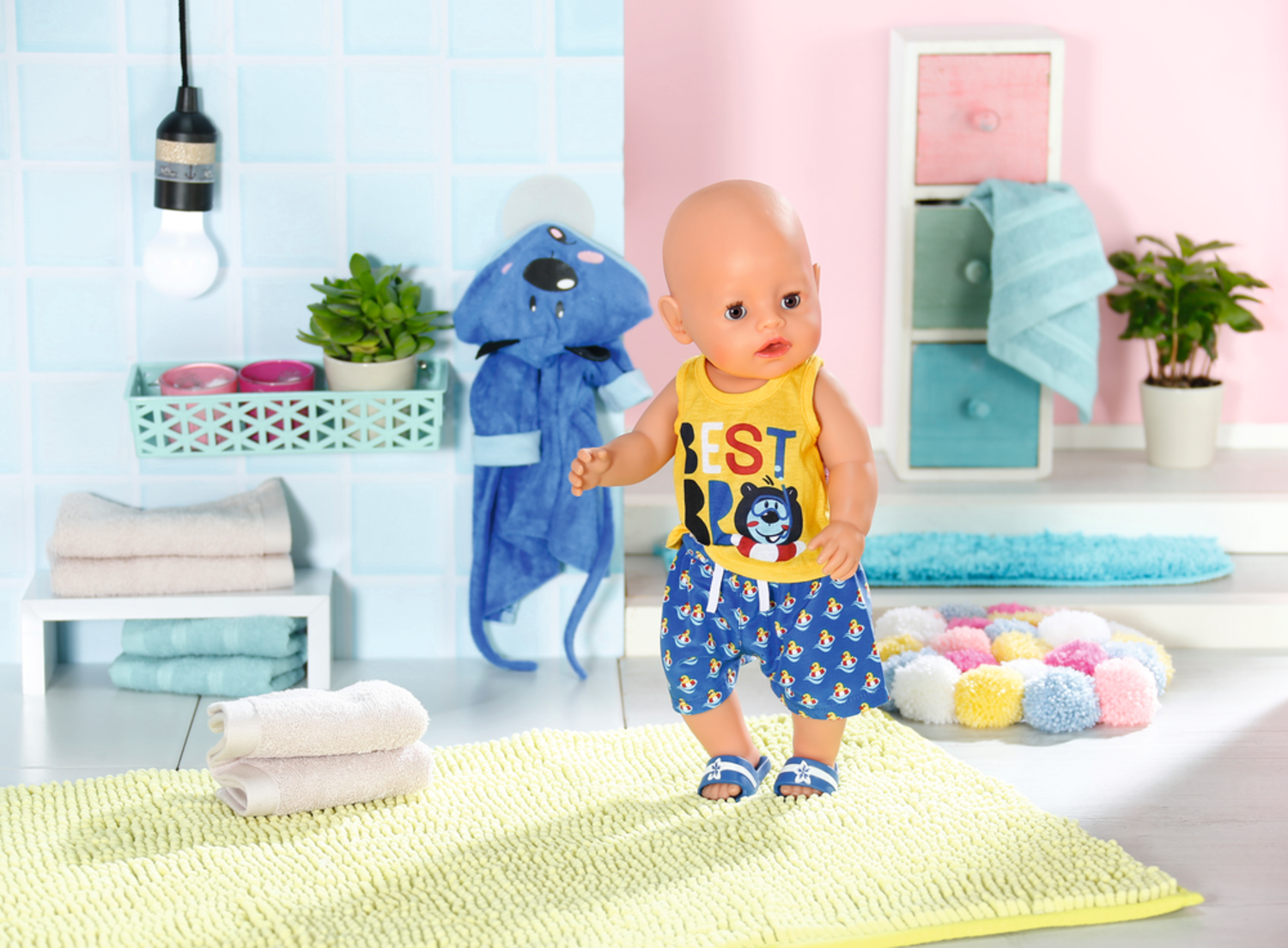 Lalka dla dziecka pokazana jest w łazience, w żółtej koszuli i niebieskich spodenkach. Jest ręcznik i szlafrok.