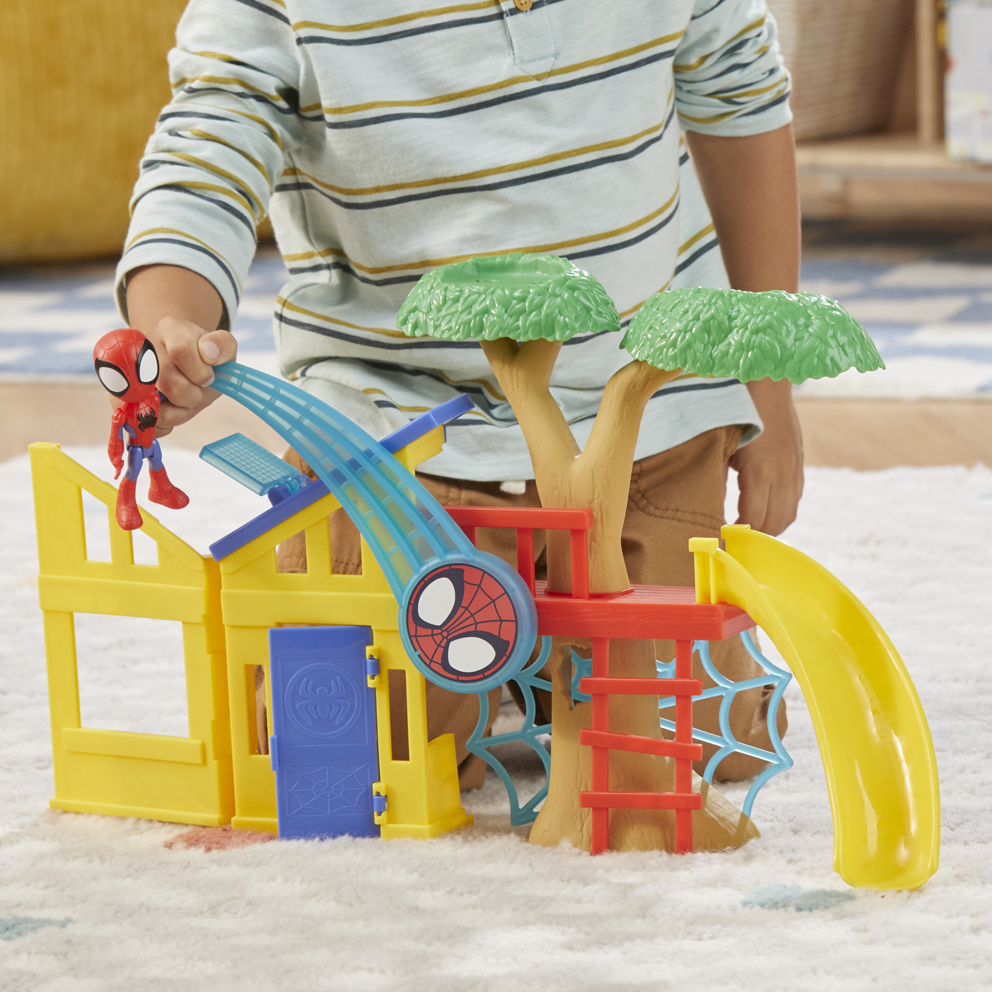 Un bambino gioca con un playset di Spider-Man. Ha una casa gialla, un albero, uno scivolo e una figura di Spider-Man.
