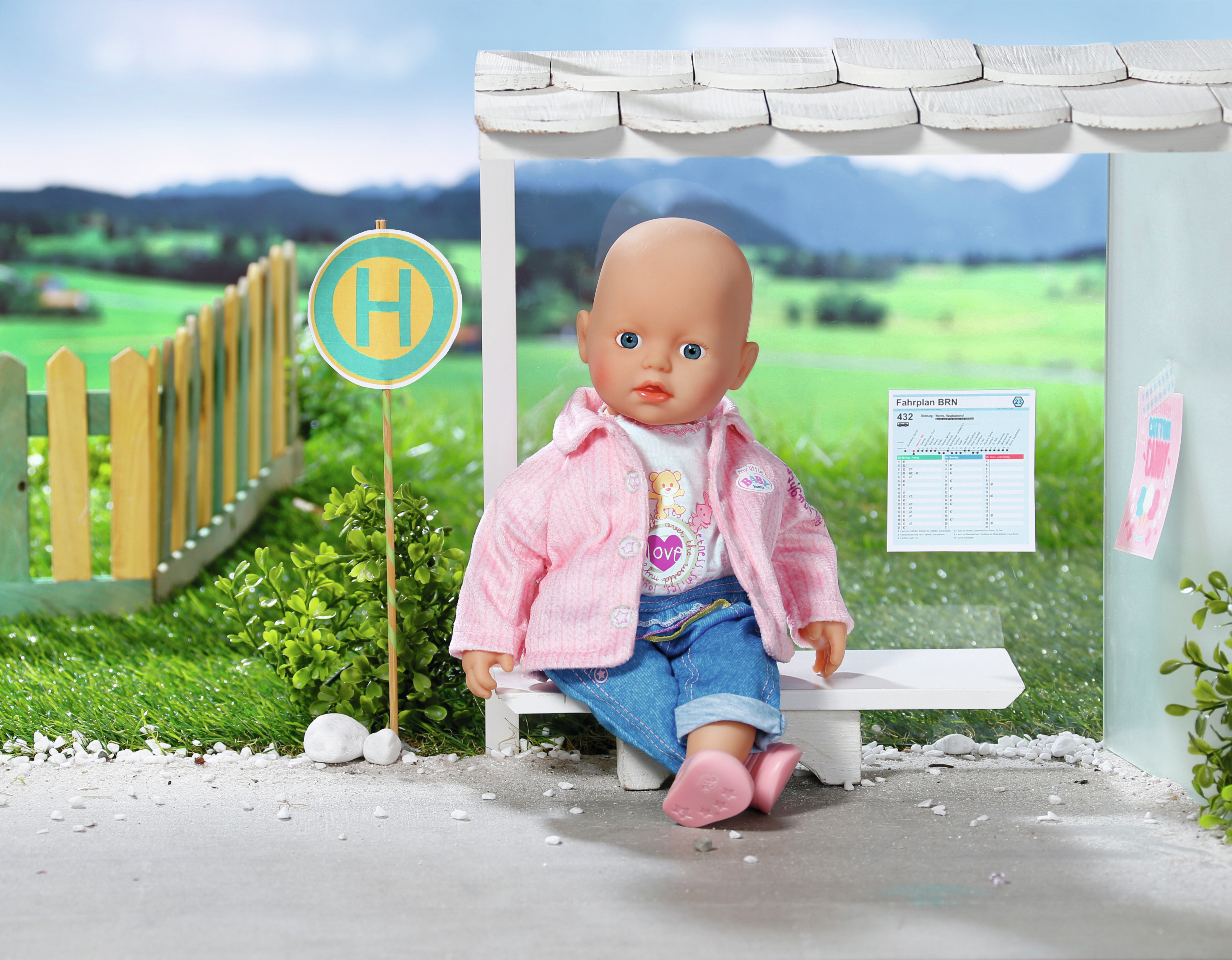 Lalka na przystanku autobusowym ze znakiem. Lalka ma na sobie różową kurtkę, dżinsy i różowe buty.