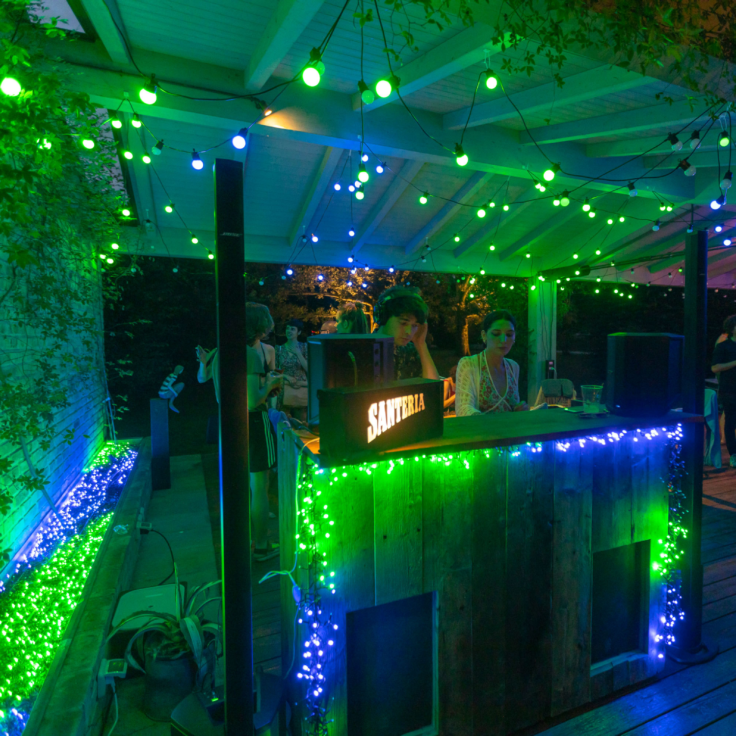 Bar na świeżym powietrzu z zielonymi i niebieskimi światłami. Dwie osoby za barem. Zielone liście w tle.