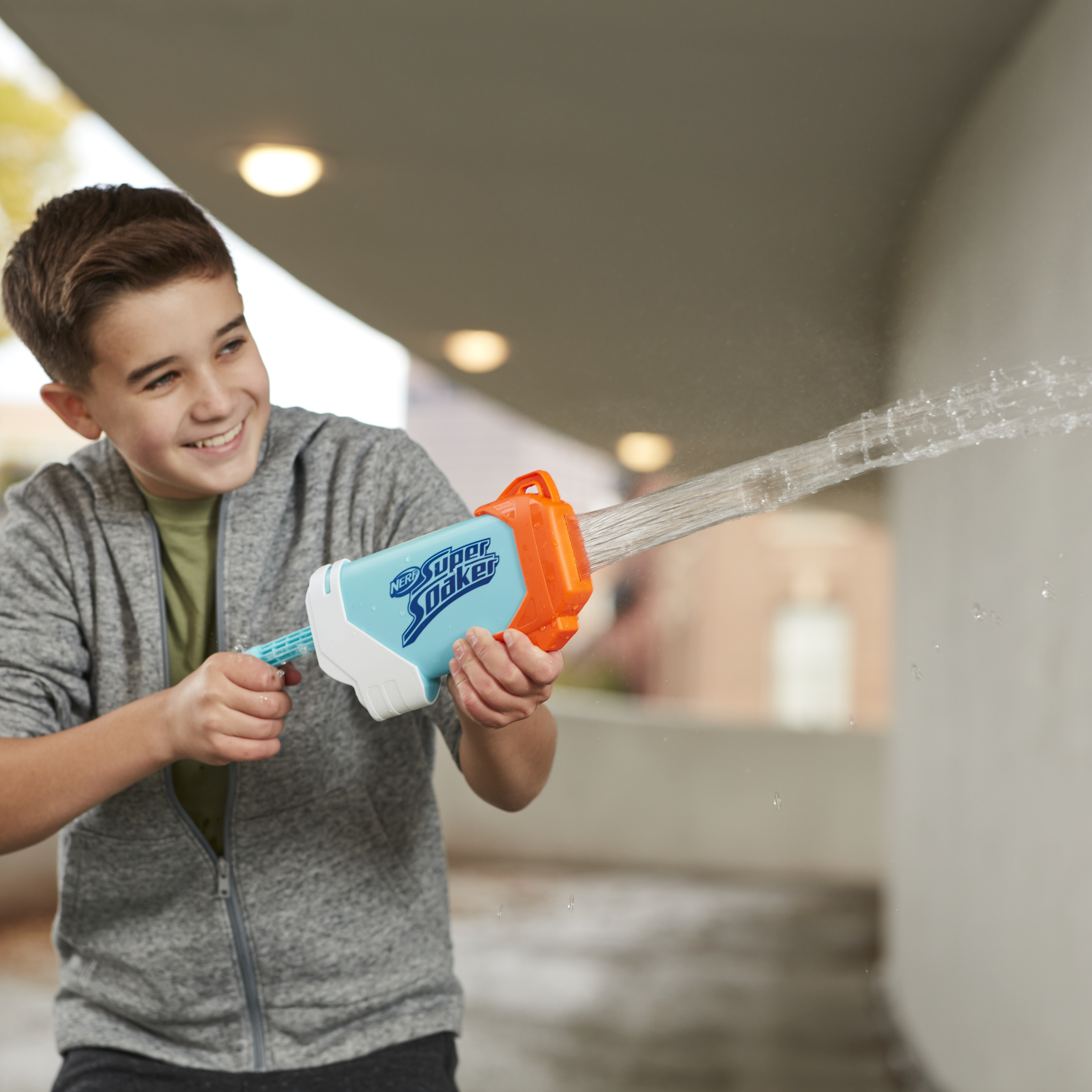 Niño sonriendo, sosteniendo una pistola de agua Super Soaker azul y naranja, rociando agua.