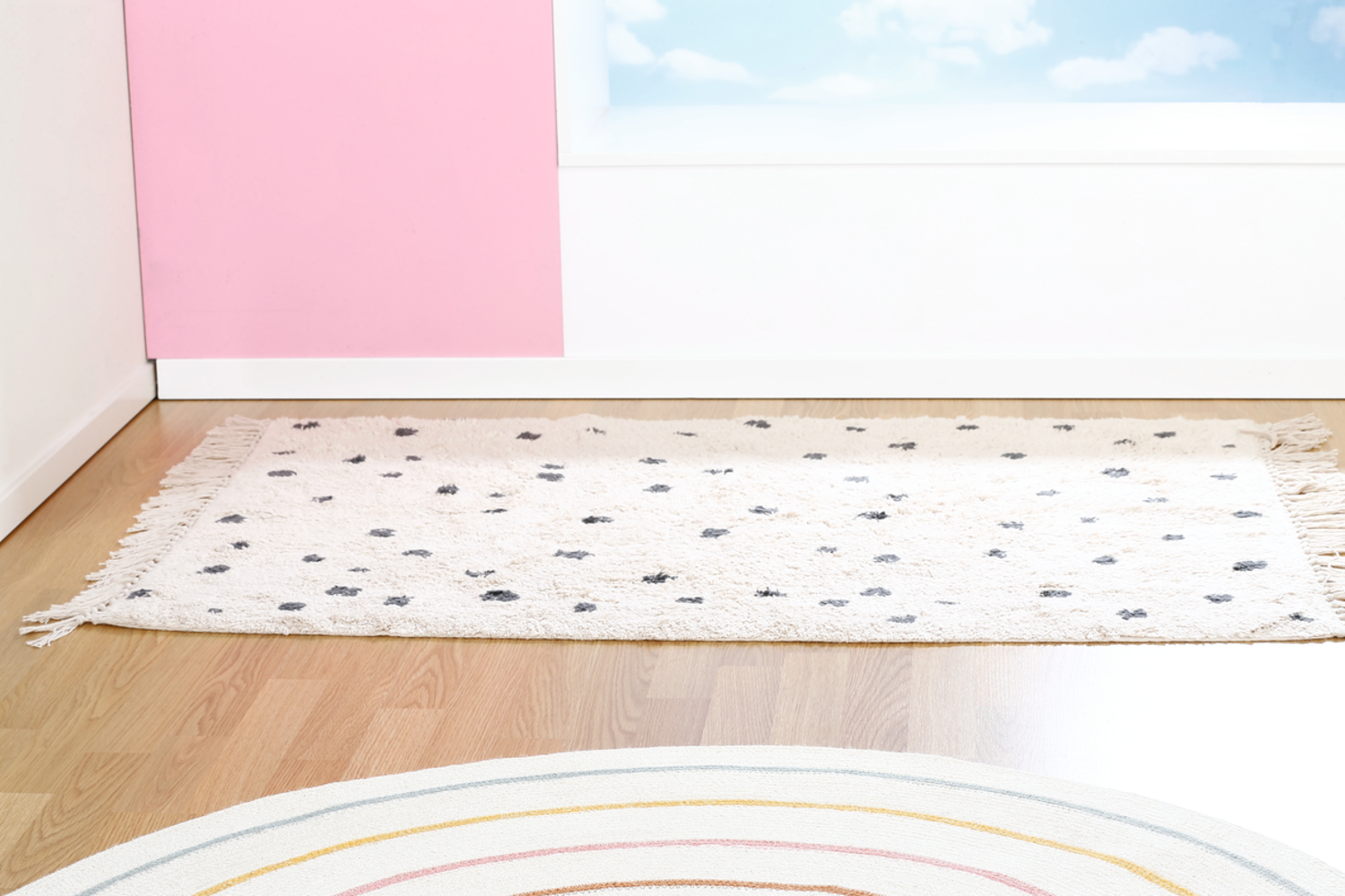 Biały dywan z szarymi kształtami chmur na drewnianej podłodze. Różowa ściana po lewej, biała ściana z niebieskim niebem w tle.