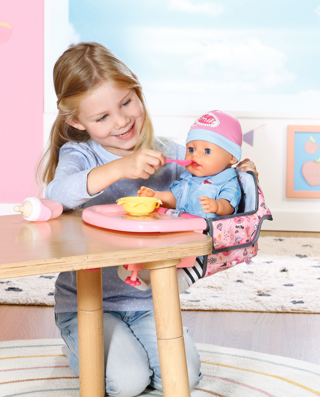 Dziewczynka karmi lalkę w krzesełku do karmienia. Dziewczynka się uśmiecha. Lalka ma różową czapeczkę i niebieskie oczy. Żółta miska i butelka.
