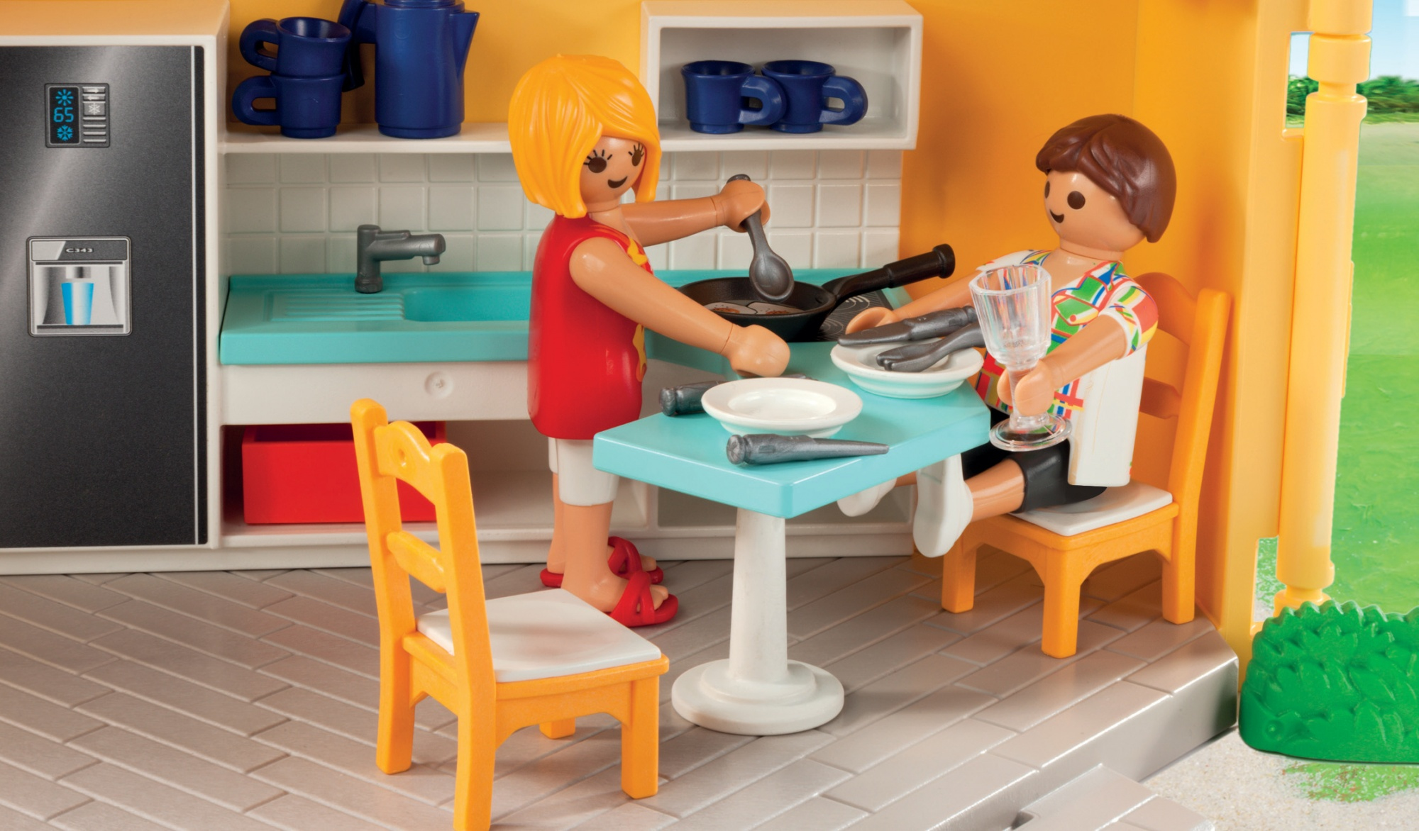 Escena de cocina Playmobil. Dos figuras, una cocinando, la otra sentada en la mesa. Colores brillantes, estilo caricatura.