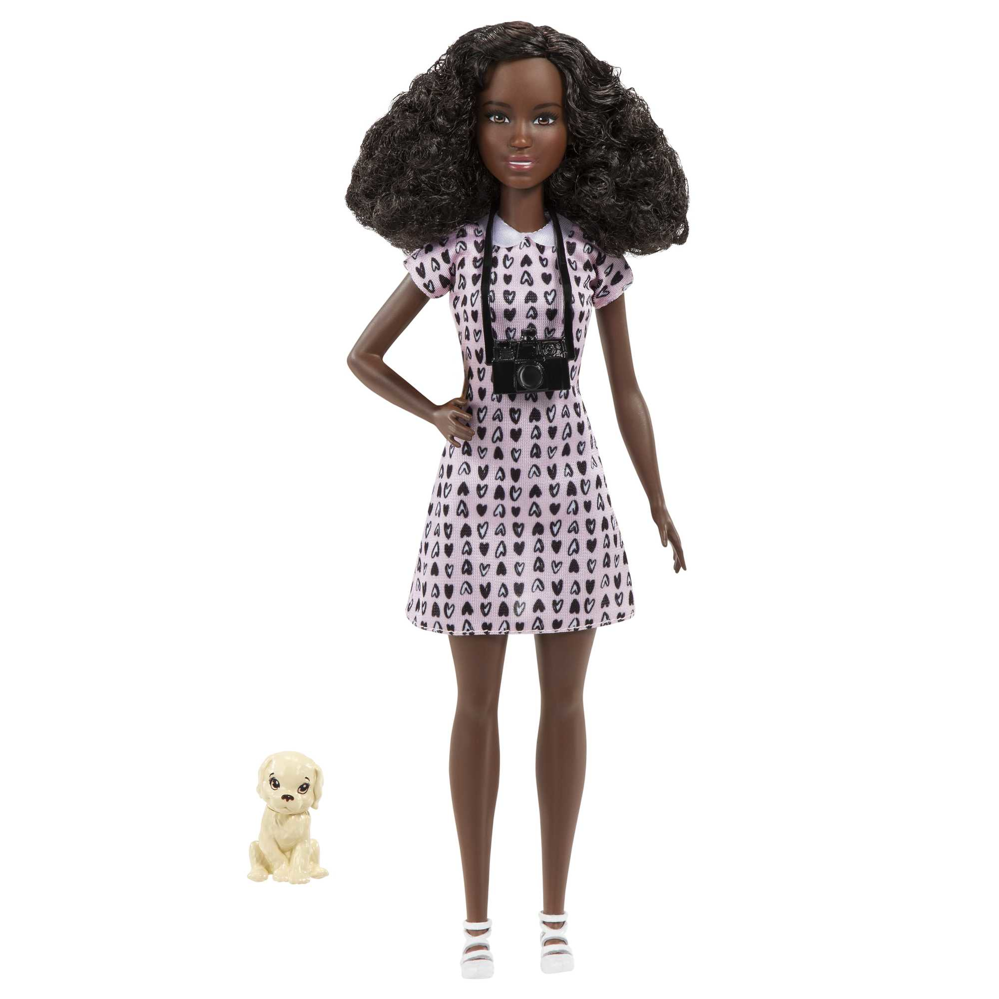Eine schwarze Barbie-Puppe in einem herzförmigen Kleid, die eine Kamera hält, mit einem kleinen Hund.