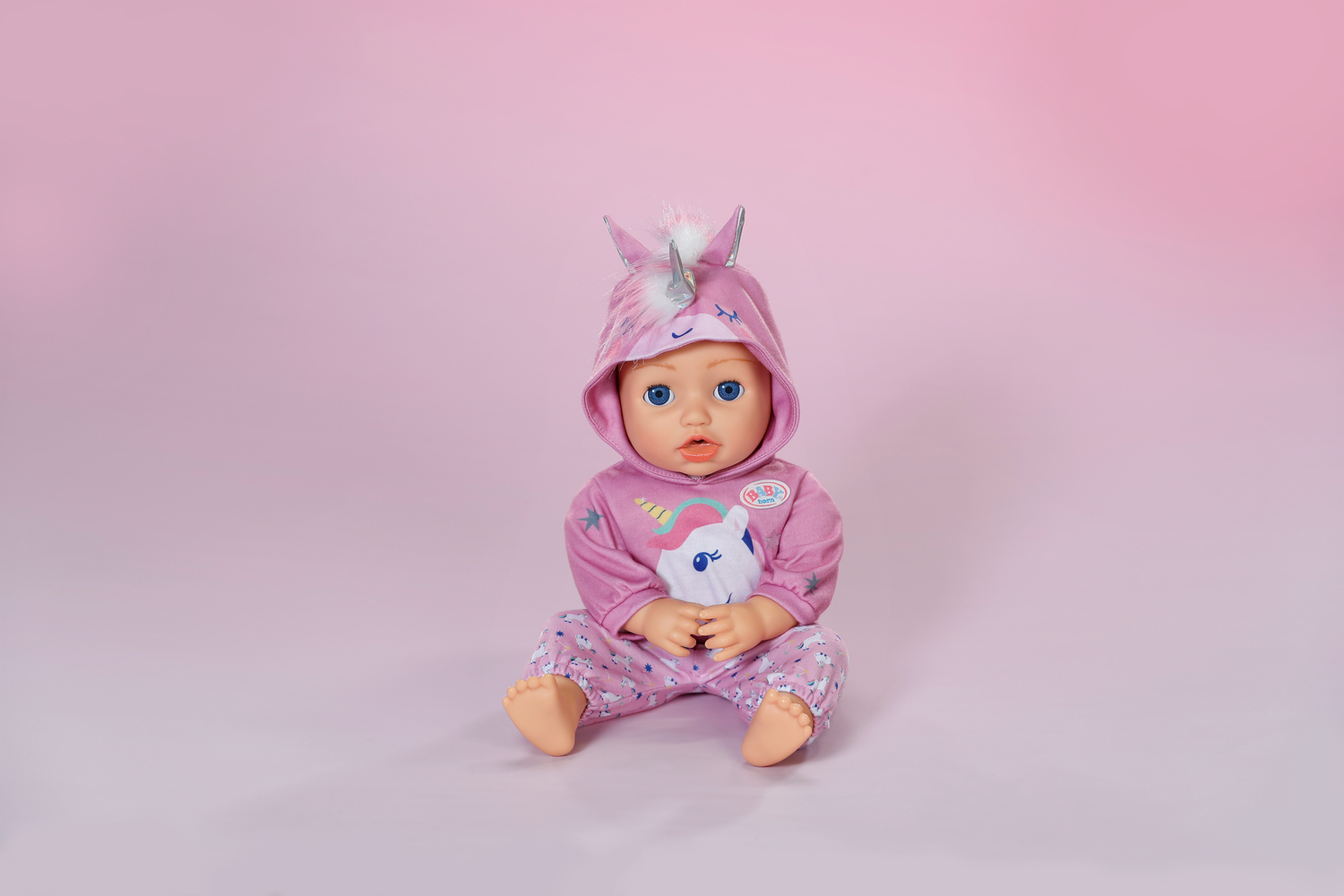 Lalka w różowej piżamie z jednorożcem, siedząca na różowym tle.