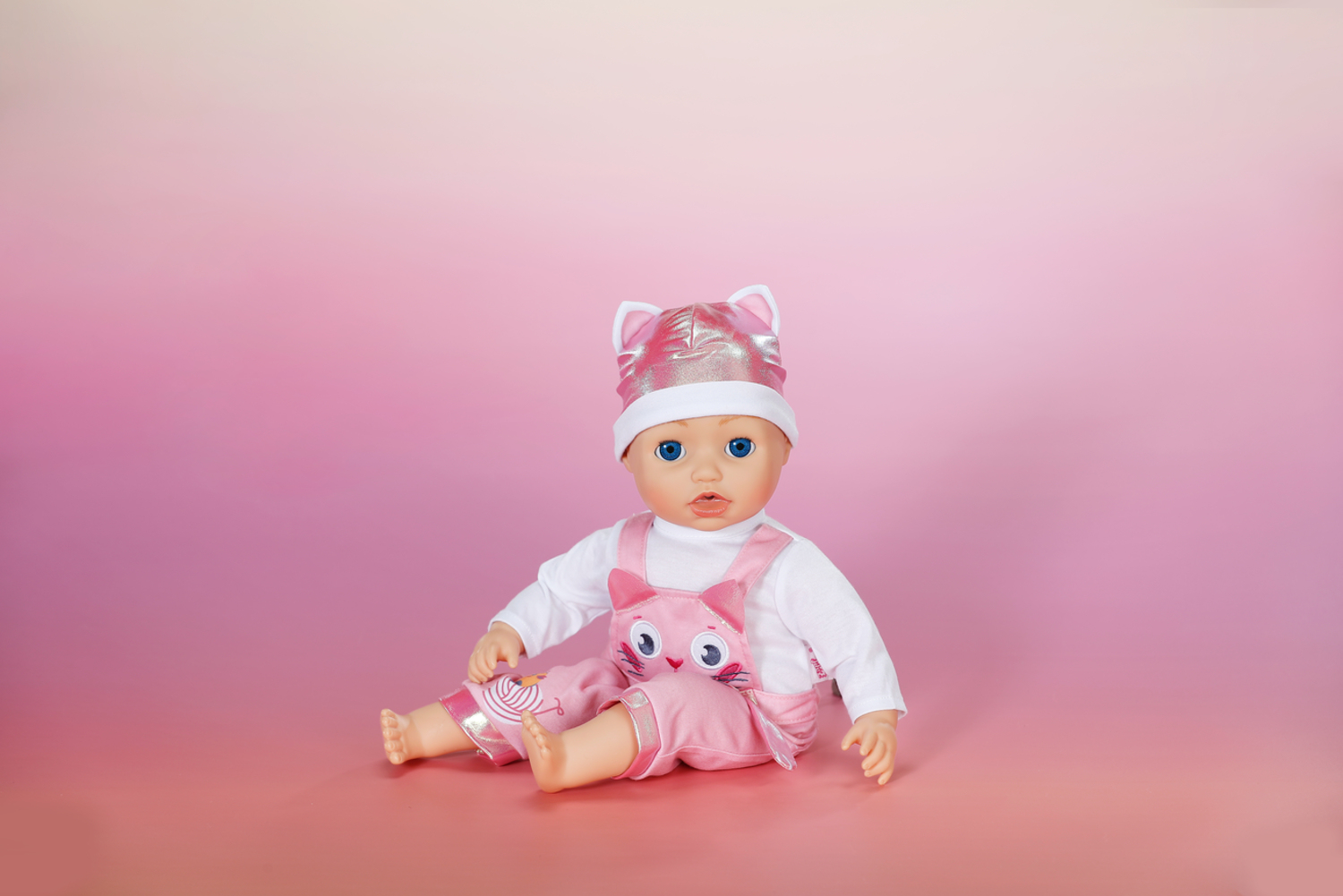 Lalka w różowym stroju z motywem kota, siedząca na różowym tle z gradientem.