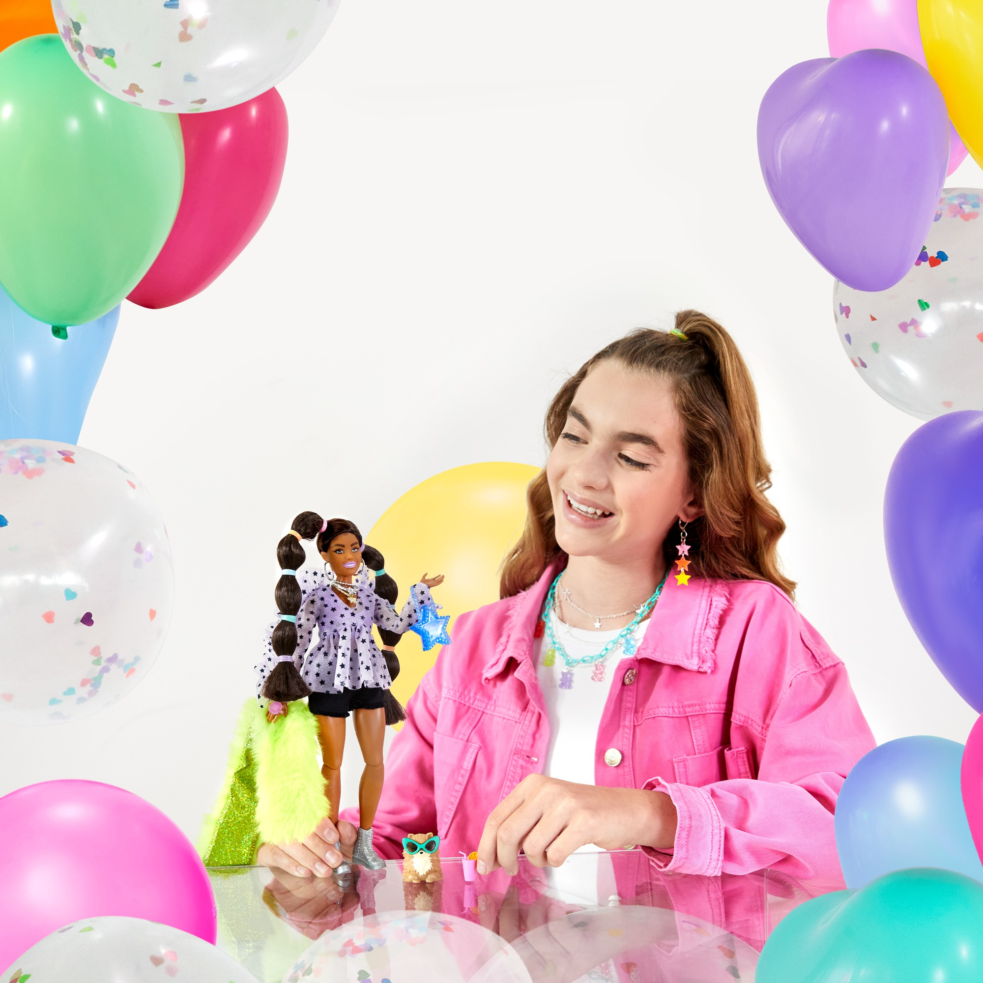 Ein Mädchen lächelt und hält eine Barbie-Puppe, umgeben von Ballons; die Puppe hat Zöpfe.