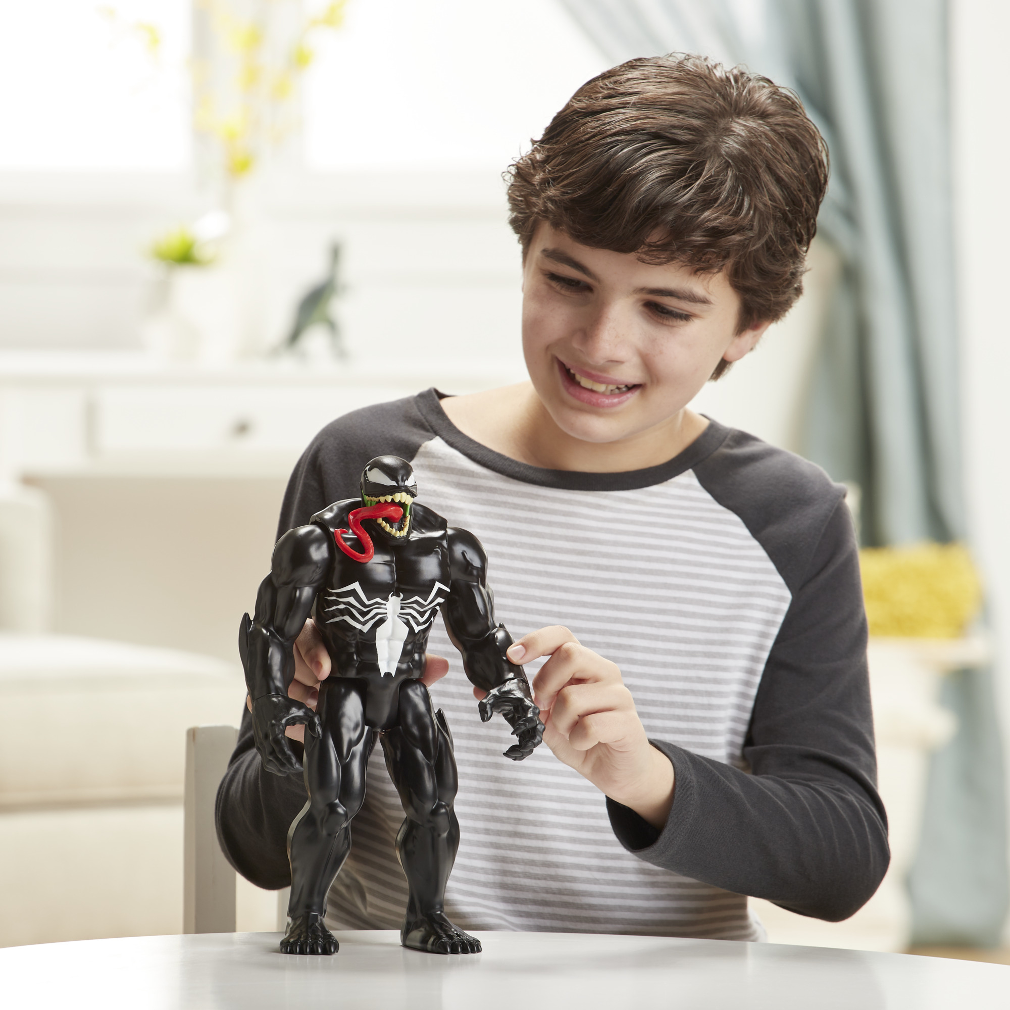 Ragazzo sorridente che gioca con un action figure di Venom. Tavolo bianco e sfondo casa.