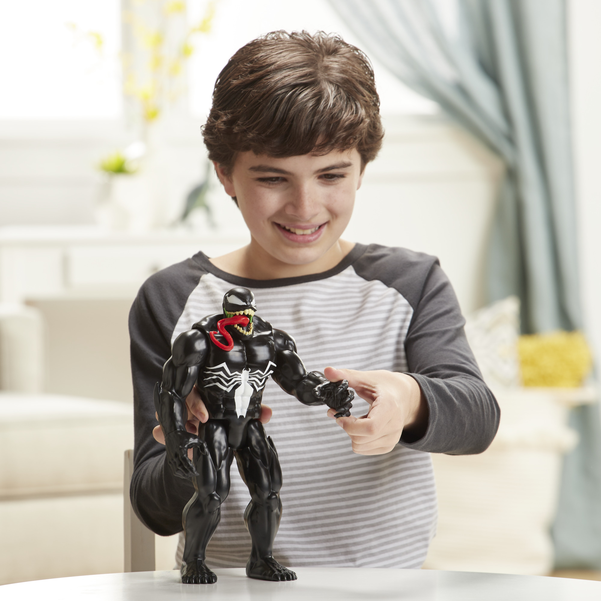 Ragazzo che tiene in mano un action figure di Venom. Il bambino sorride. Tavolo bianco e sfondo casa.