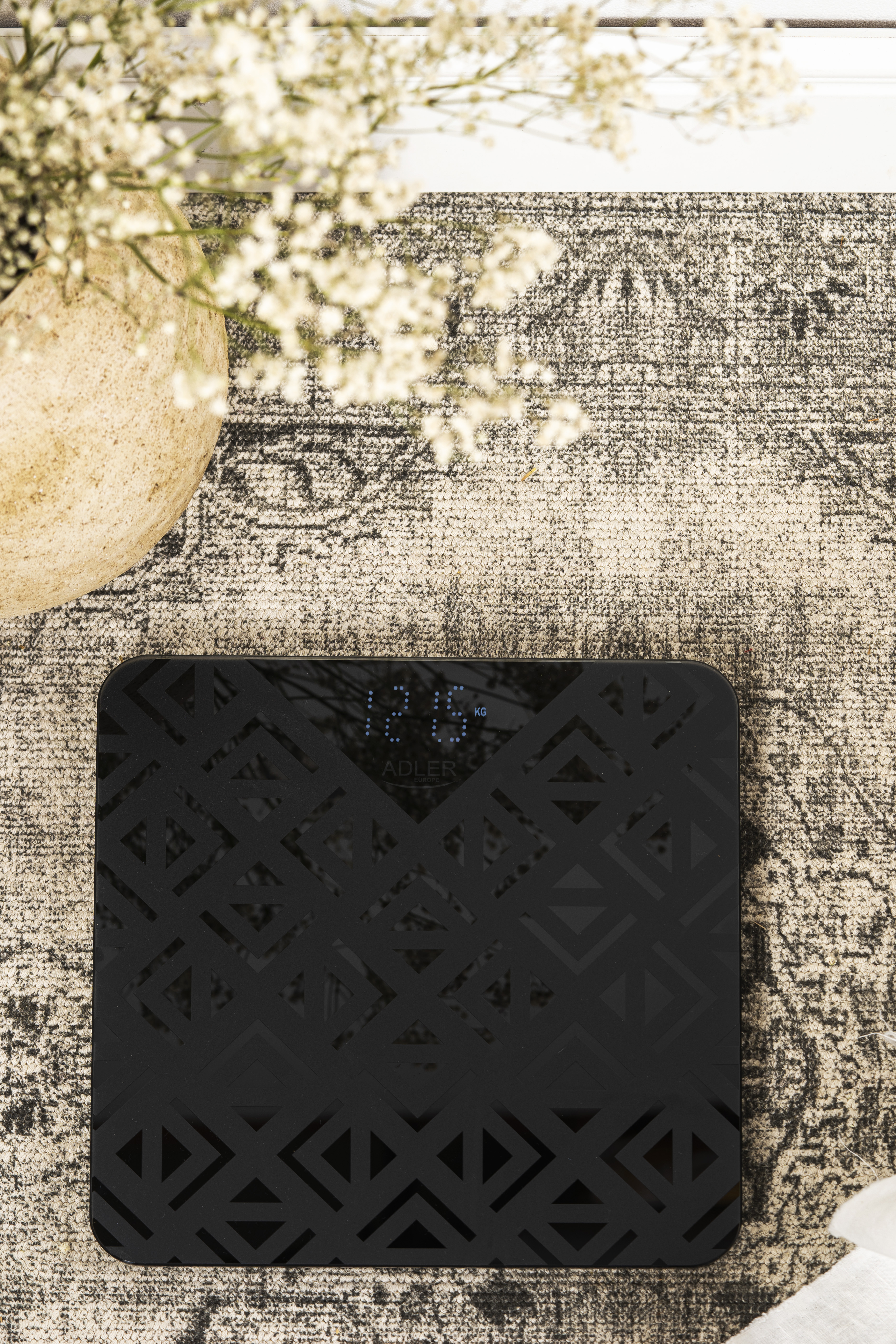 Czarna waga łazienkowa z wzorzystą powierzchnią, pokazująca 12,6 kg. W tle wazon i kwiaty.
