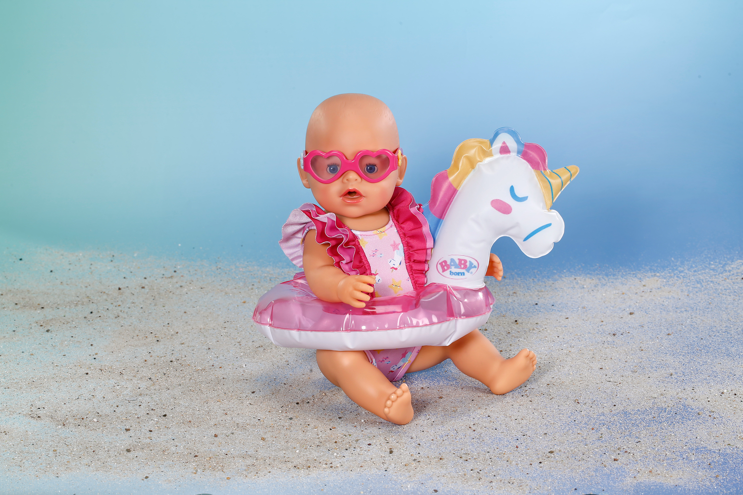 Lalka w okularach przeciwsłonecznych, kostiumie kąpielowym i jednorożcu na piaszczystej plaży.