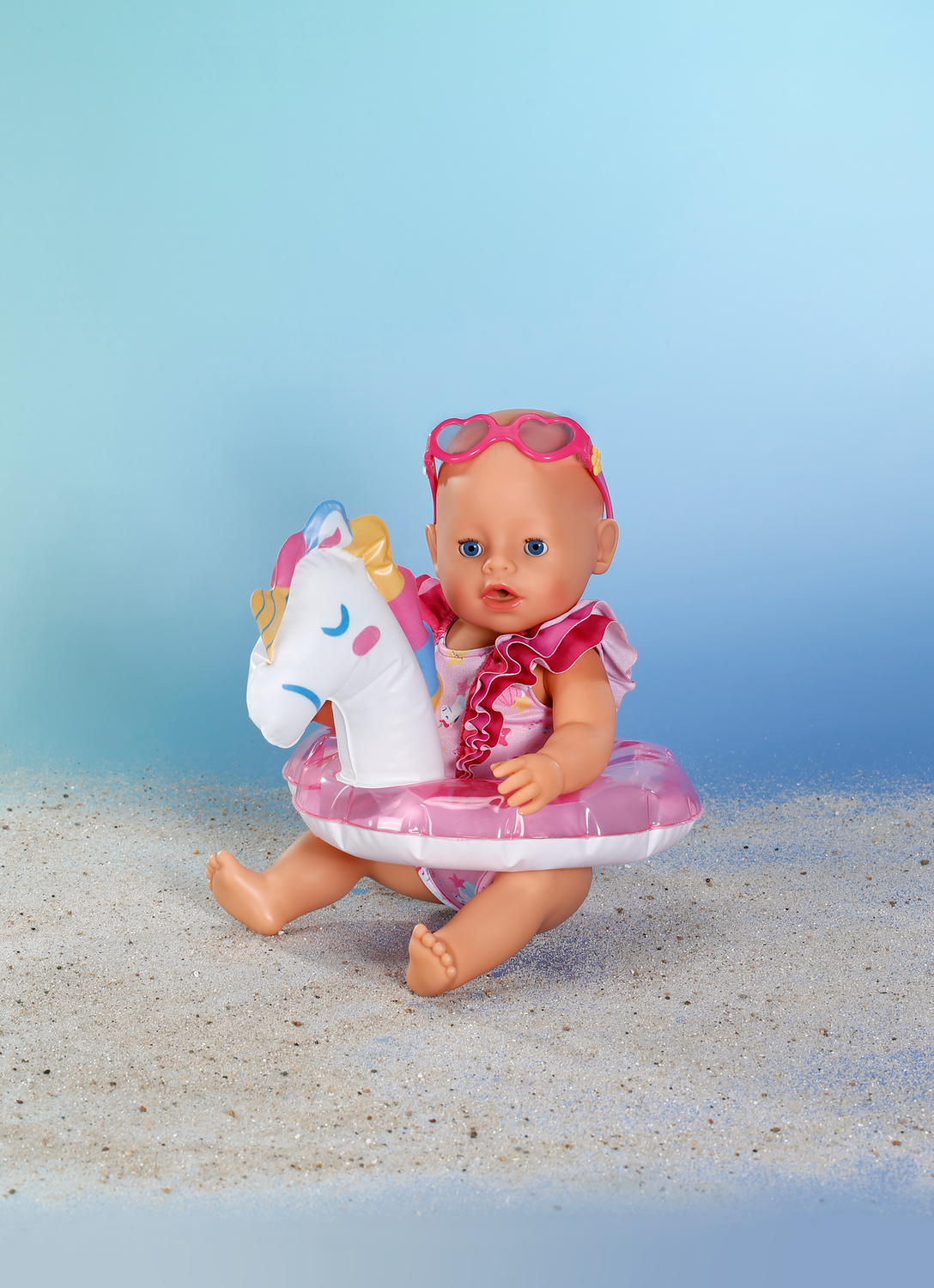 Lalka w kostiumie kąpielowym, jednorożcu i okularach przeciwsłonecznych, siedząca na plaży z niebieskim niebem.