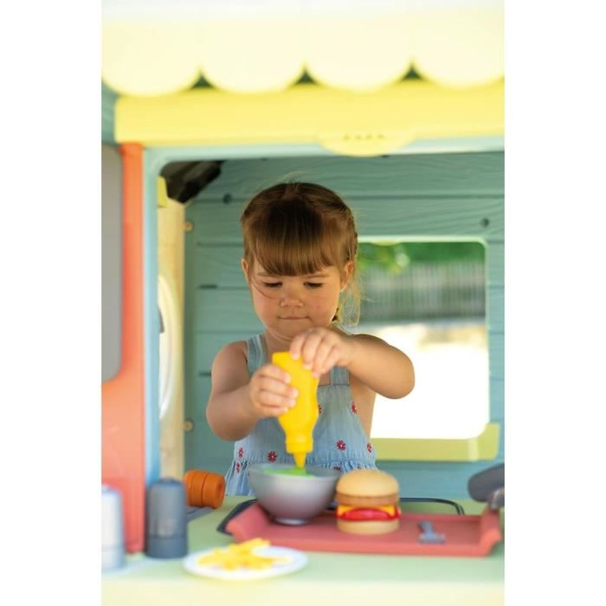 Una bambina aggiunge salsa gialla a una ciotola in un set da cucina giocattolo con un hamburger.