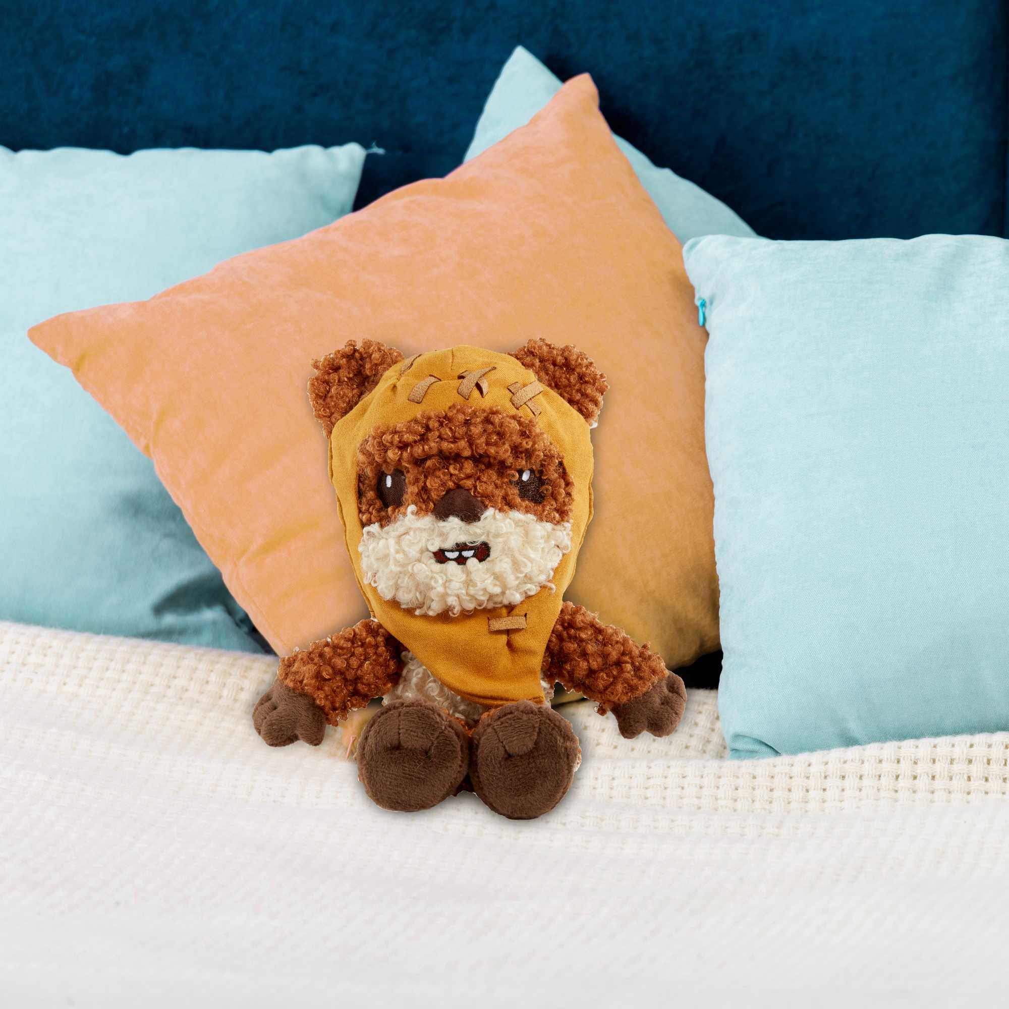 Un peluche Ewok su una coperta bianca con cuscini colorati sullo sfondo.