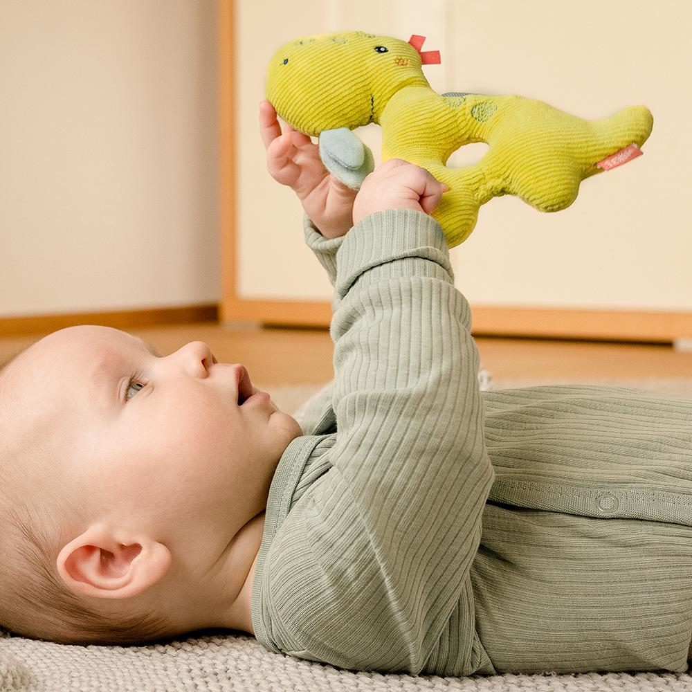 Niemowlę trzymające żółtą grzechotkę dinozaura. Niemowlę ma na sobie zielony pajac na szarej macie.