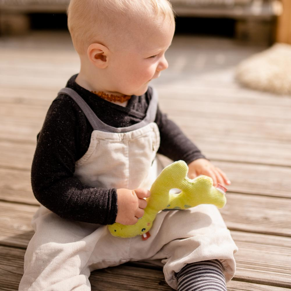 Niemowlę siedzące na drewnianej podłodze bawiące się żółtą grzechotką dinozaura.