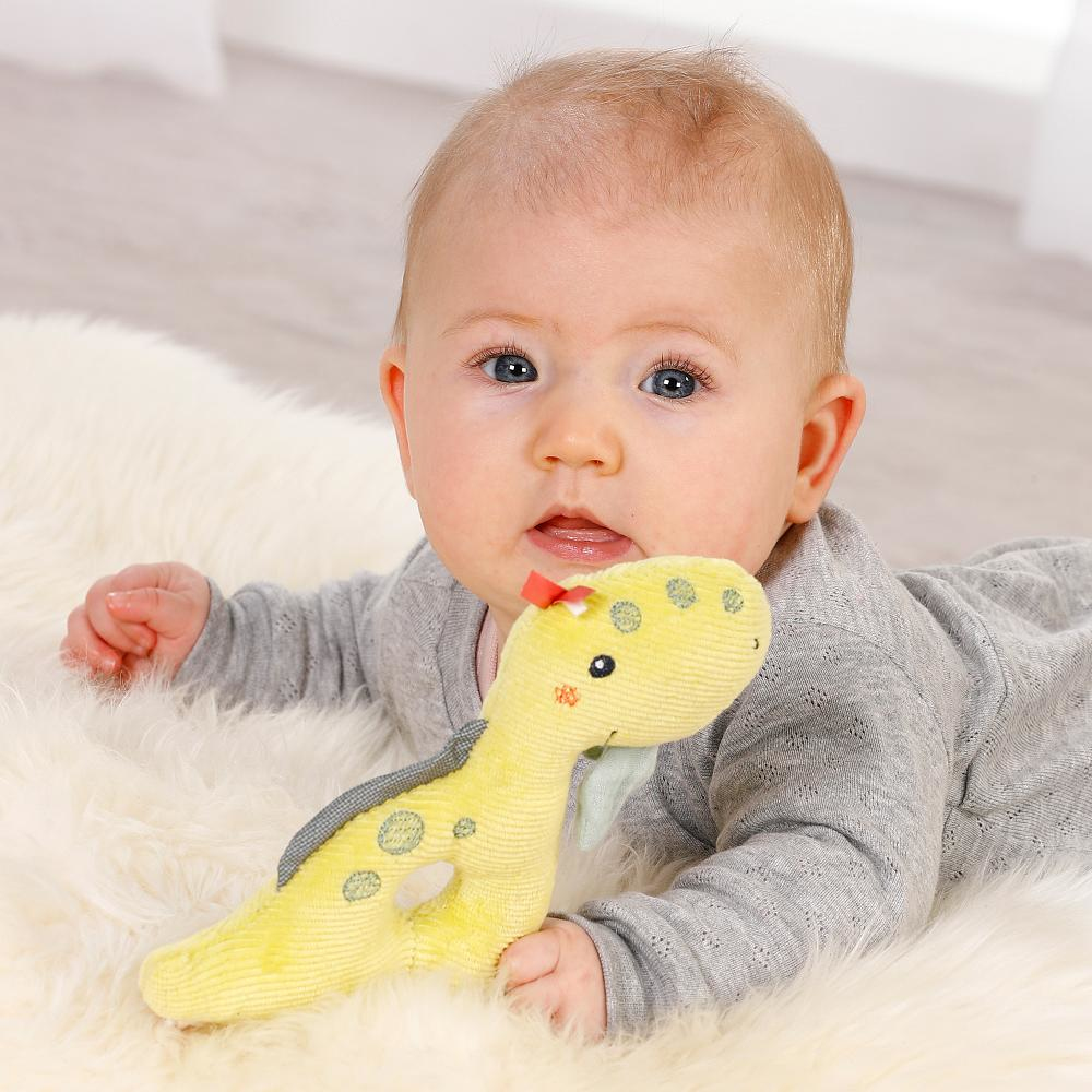 Dziecko leżące na puszystym, białym dywanie, patrzące na żółtą zabawkę-grzechotkę w kształcie dinozaura.