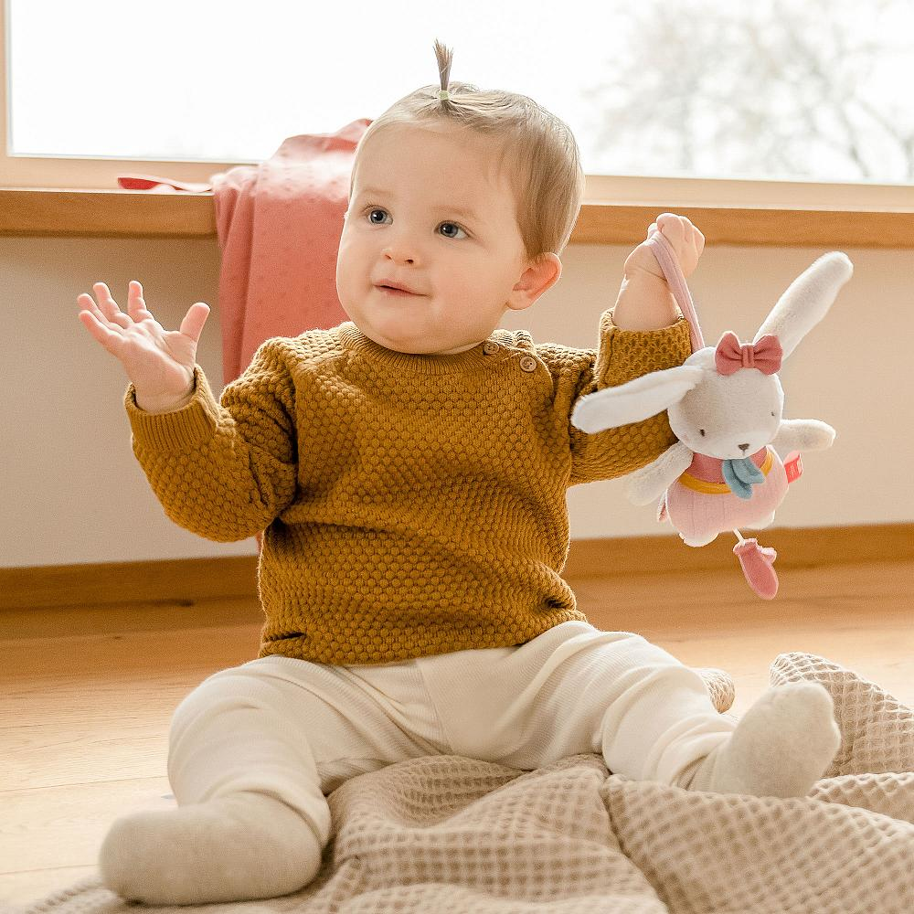 Baby sitzt auf einer Decke und hält ein Plüsch-Kaninchenspielzeug. Sonniger Hintergrund.