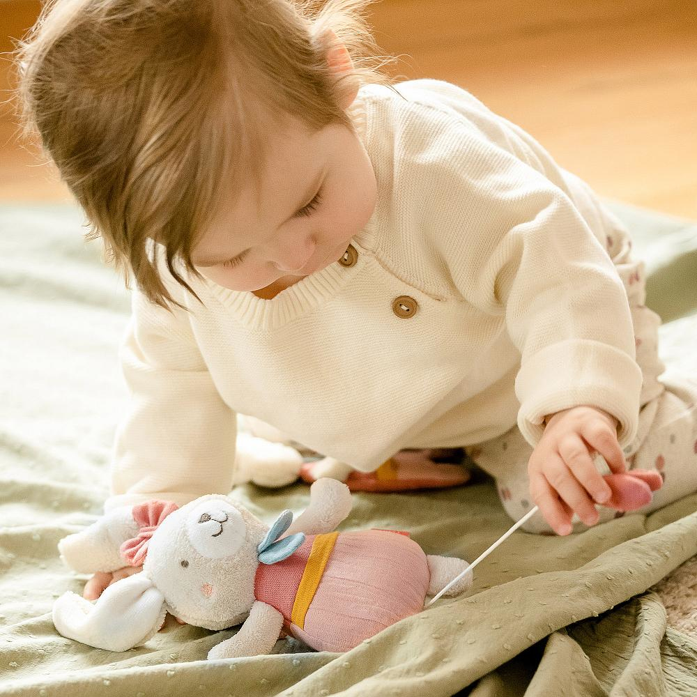 Baby spielt mit einem Spielzeug-Hasen auf einer grünen Decke. Das Baby trägt einen weißen Pullover.