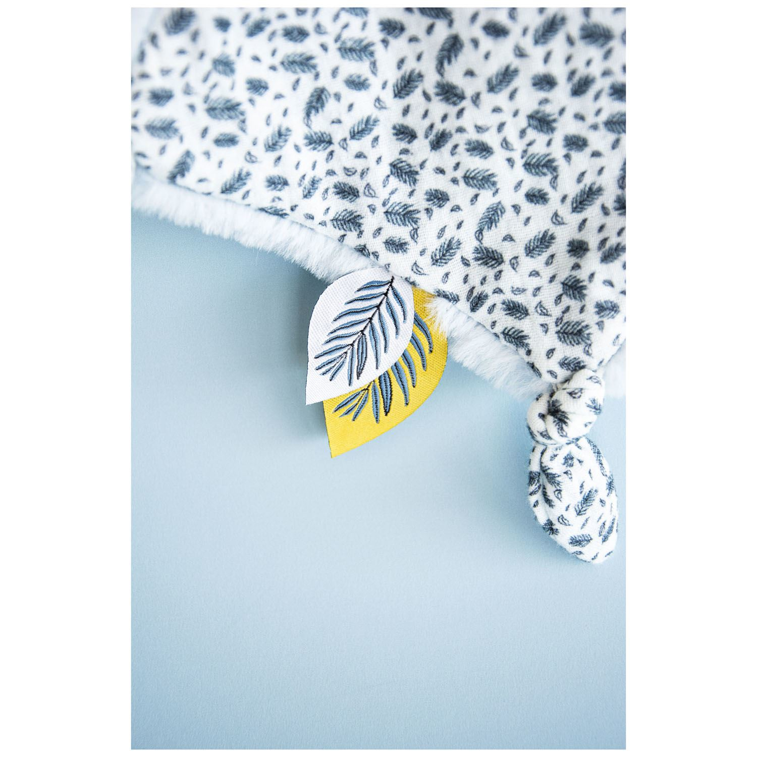 Wzorzysty koc z motywami liści oraz żółto-niebieska etykieta w kształcie liścia. Tło jest jasnoniebieskie.