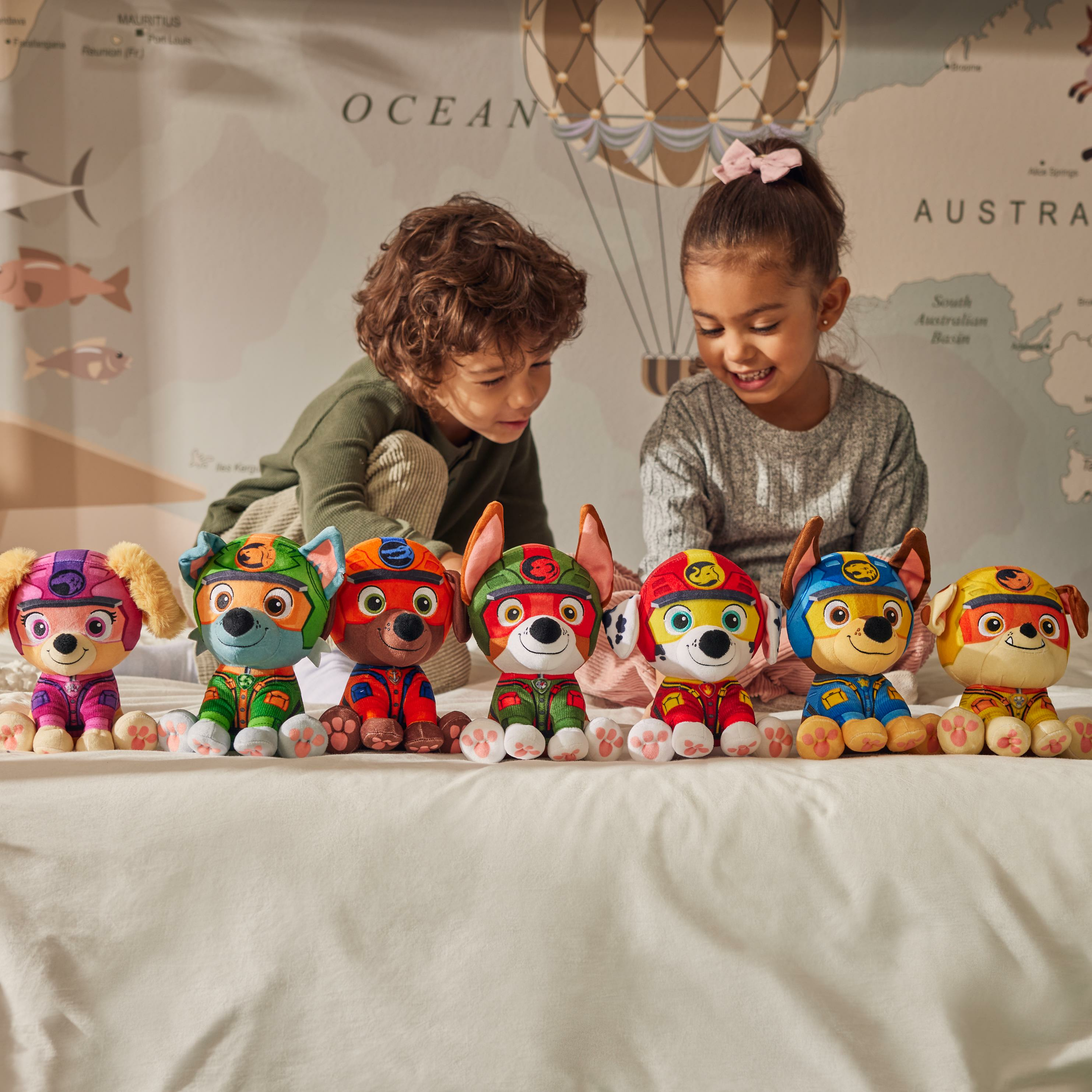 Zwei Kinder spielen mit mehreren Paw Patrol-Plüschtieren auf einem Bett mit einer Landkartenkulisse.
