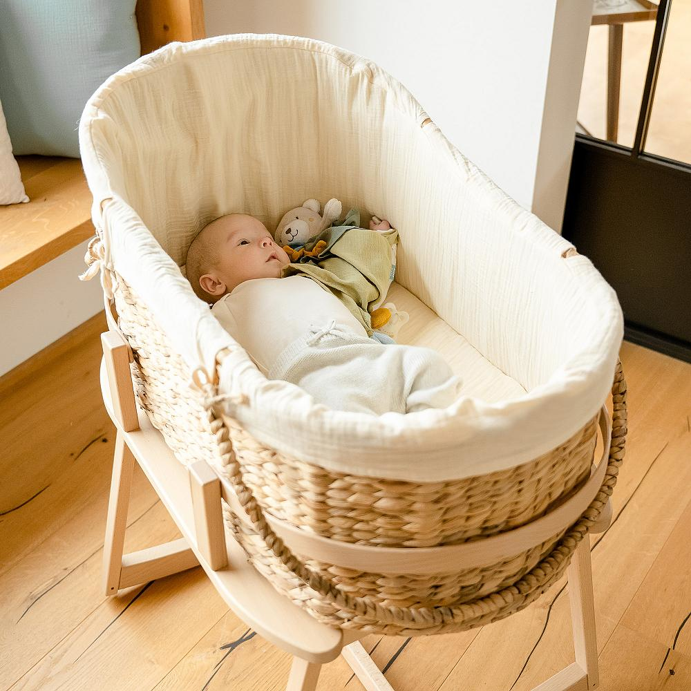 Dziecko leżące w wiklinowym koszu, z pluszową zabawką. Drewniana podłoga i okno w tle.