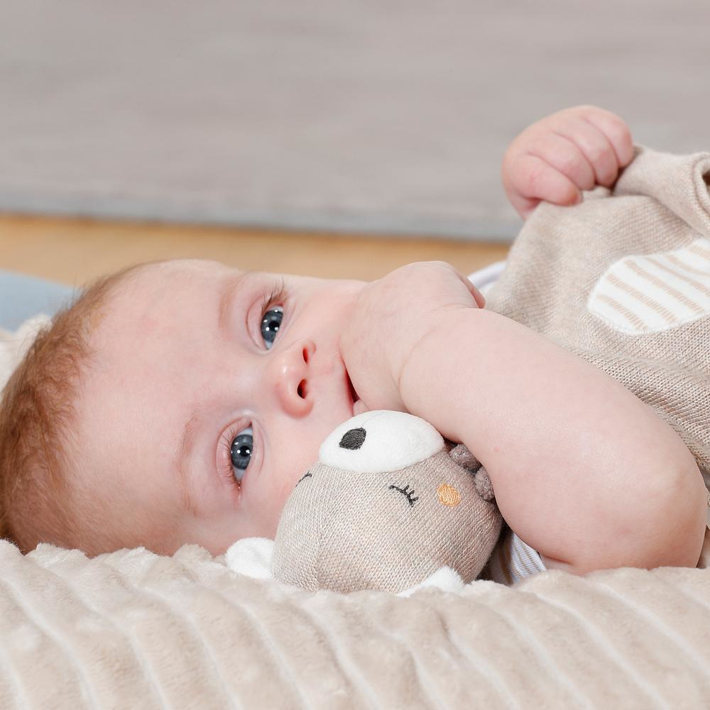 Un bambino sdraiato su una coperta tiene in mano un peluche. Il bambino ha gli occhi azzurri e guarda la fotocamera.
