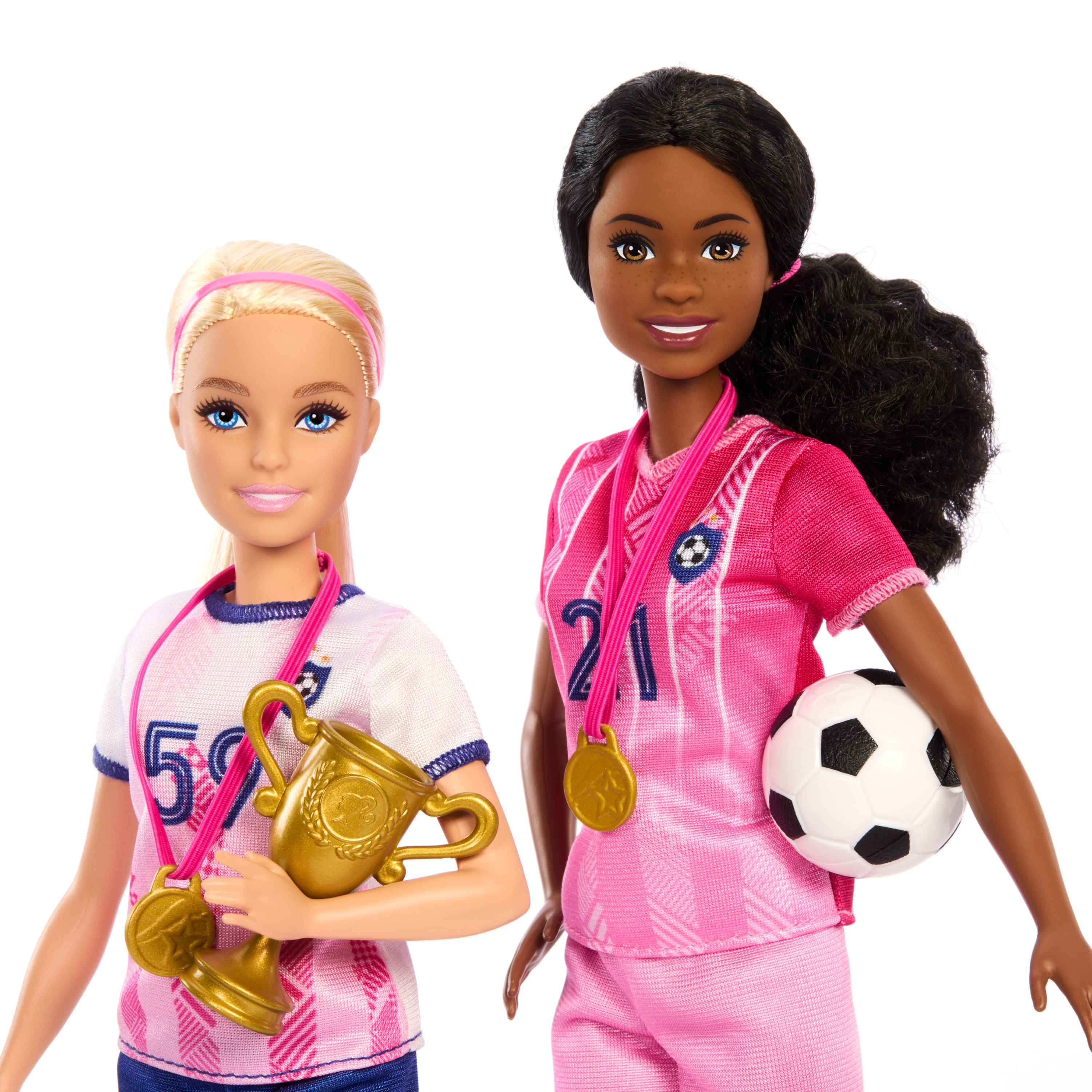 Zwei Barbie-Puppen in Fußballuniformen, eine blond, eine schwarzhaarig. Zubehör umfasst eine Trophäe, einen Ball und Medaillen.