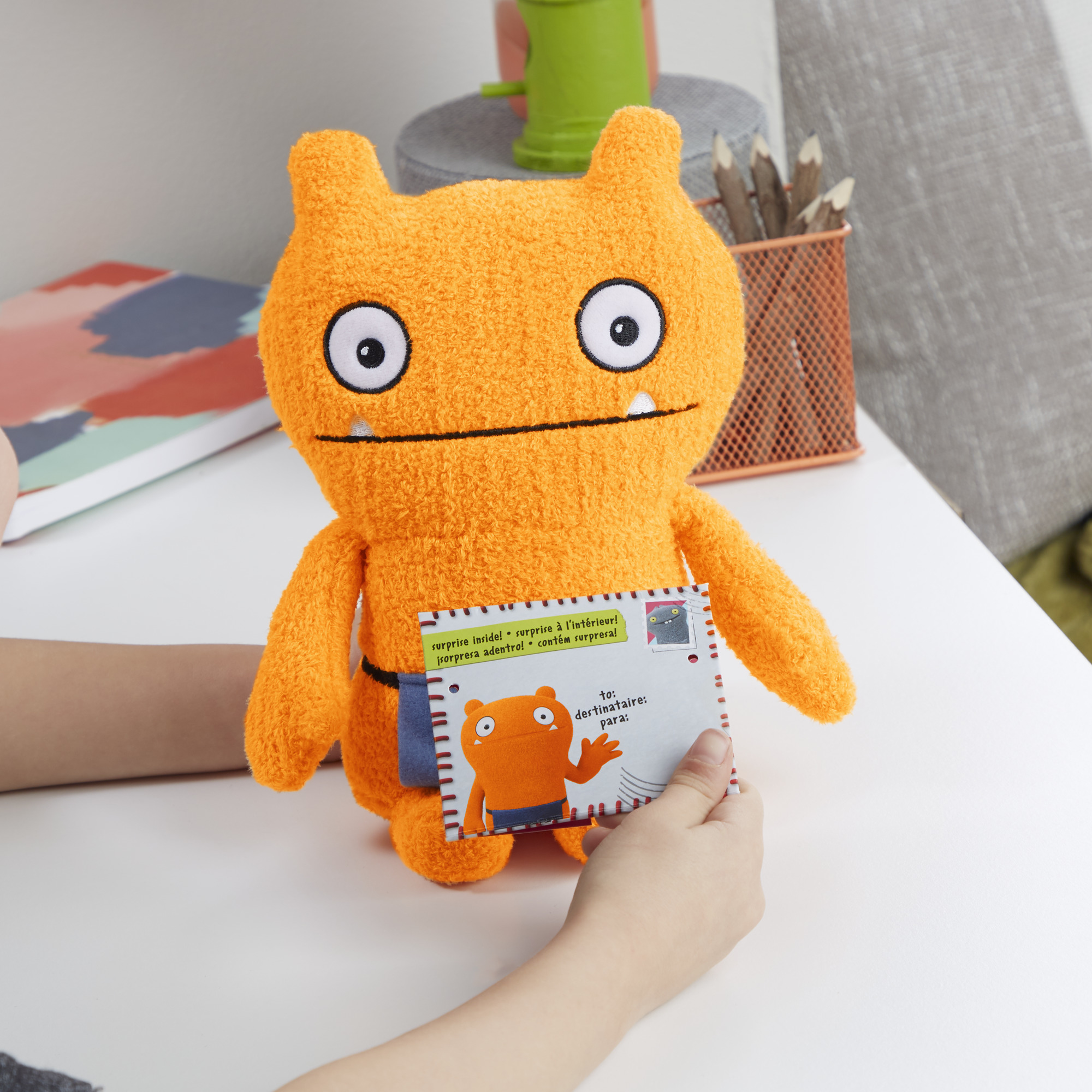 Orangenes Monster-Plüschtier hält eine Karte. Die Hände einer Person sind auf einer weißen Oberfläche zu sehen.