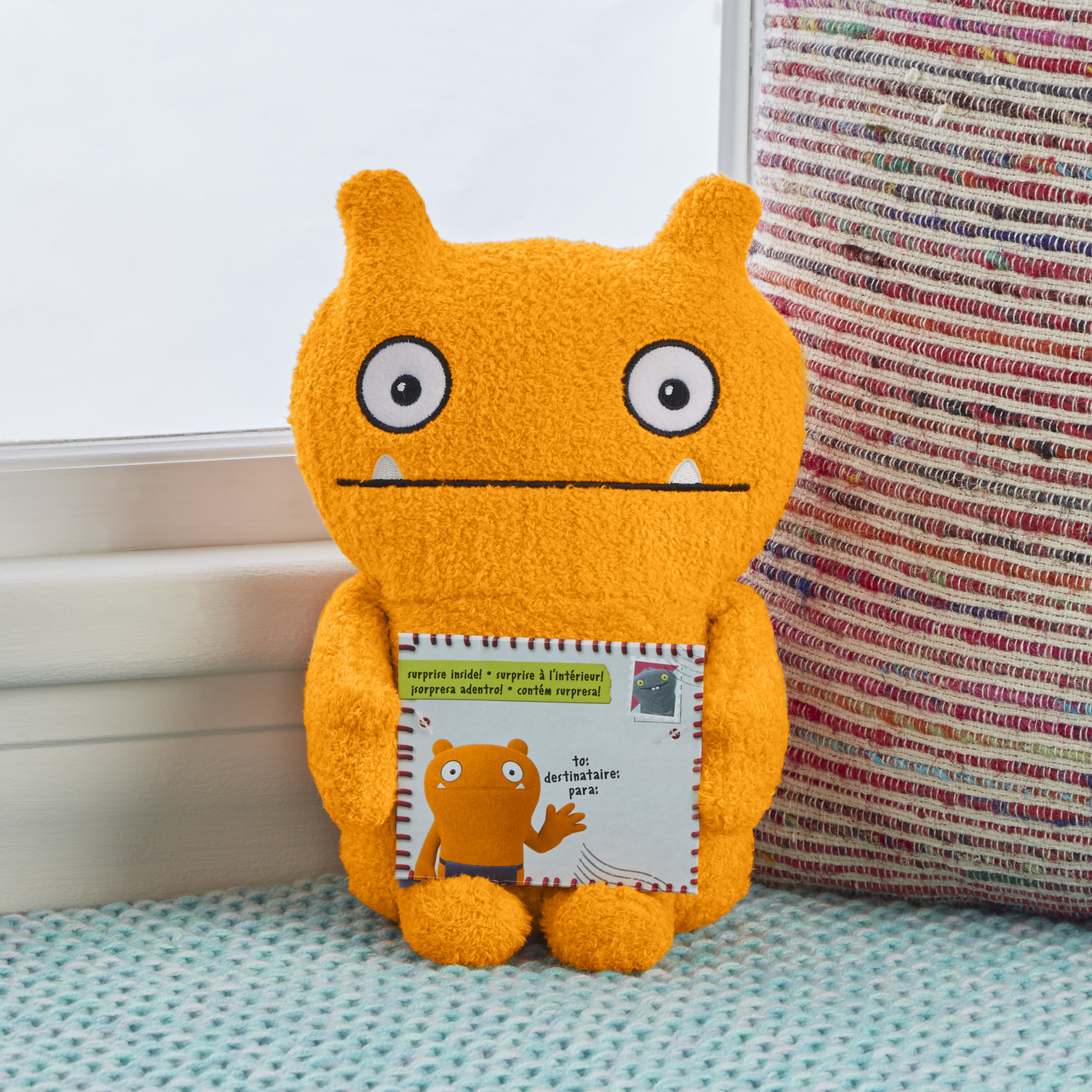 Oranges Monster-Plüschtier hält eine Karte. Es sitzt in der Nähe eines Fensters mit einem bunten Teppich.