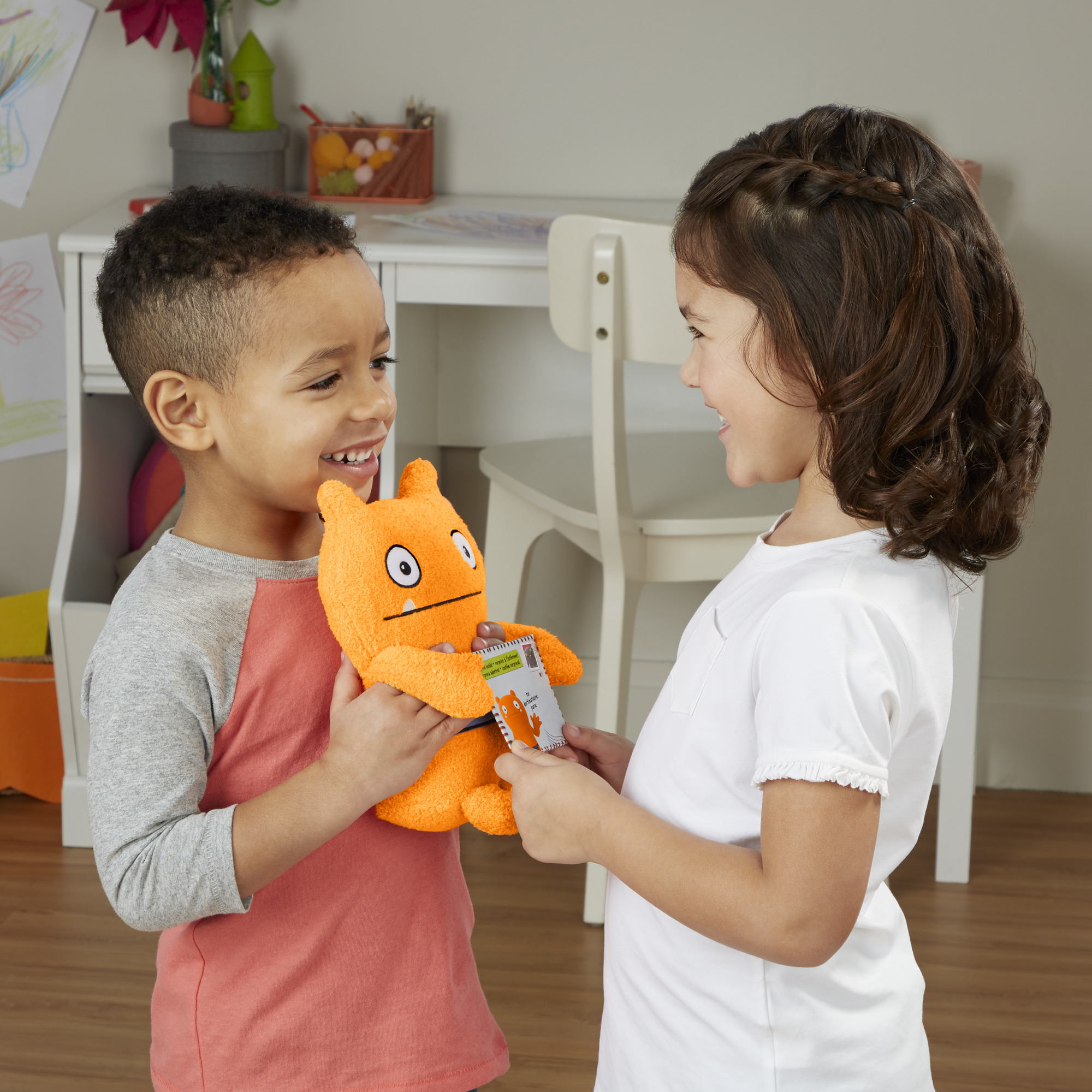 Zwei Kinder halten ein orangefarbenes Stofftier und eine Karte und lächeln sich in einem Raum an.