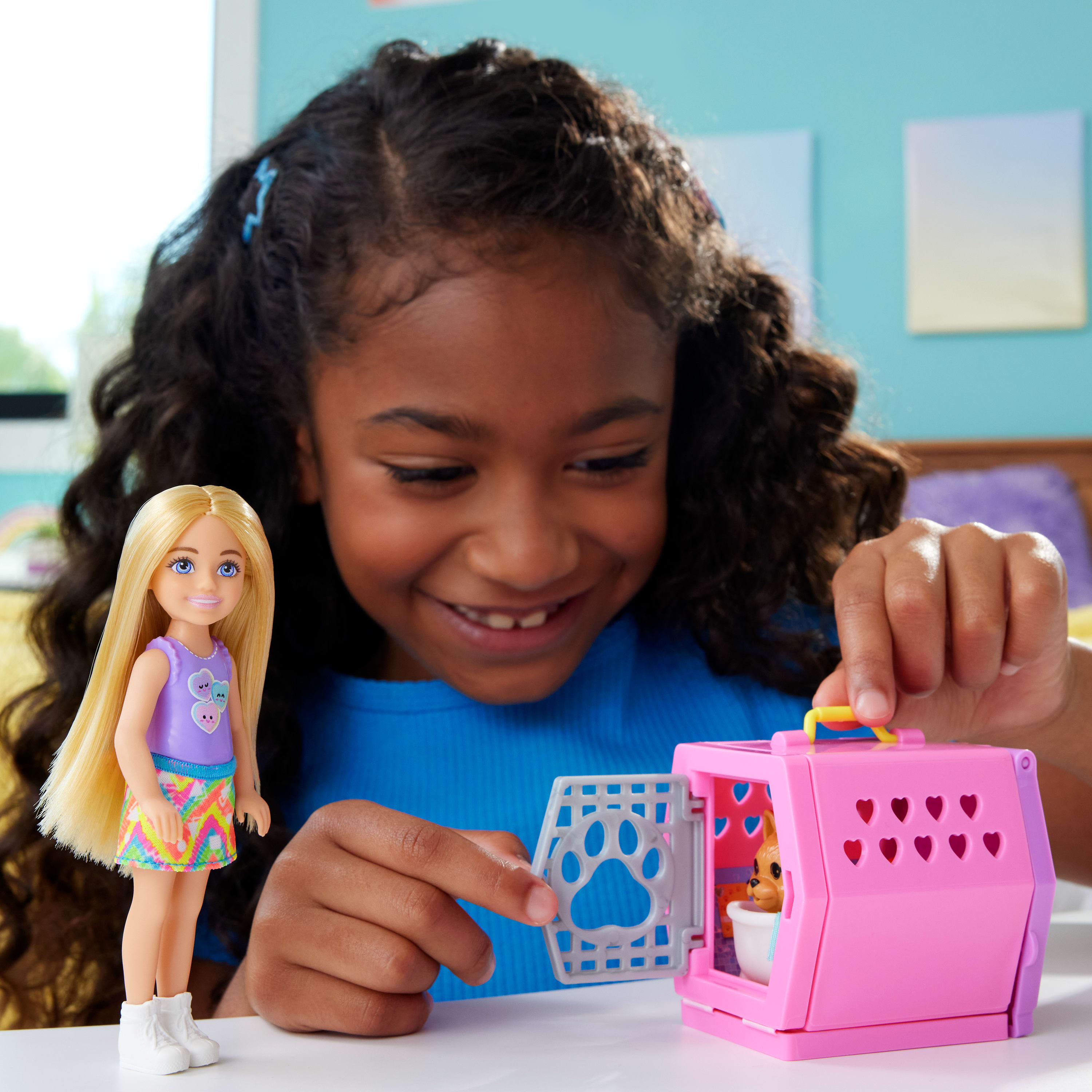 Dziewczynka uśmiecha się do lalki i różowego transportera dla zwierząt z szczeniakiem w środku, na stole.