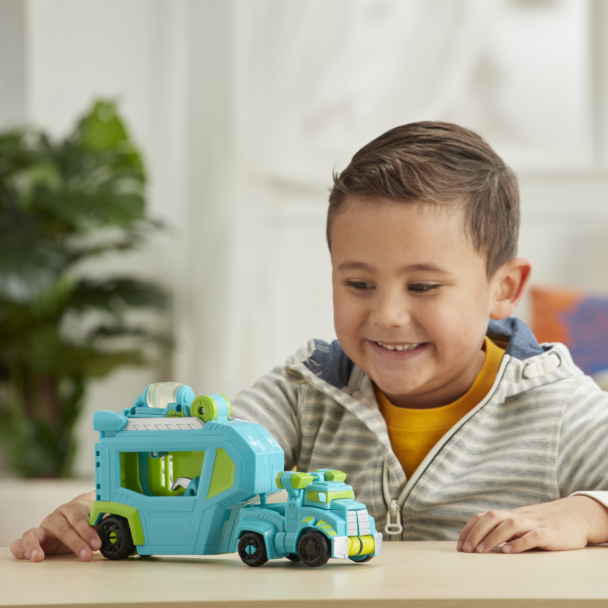 Un bambino sorride a un camion giocattolo blu e verde. È seduto su un tavolo di legno.