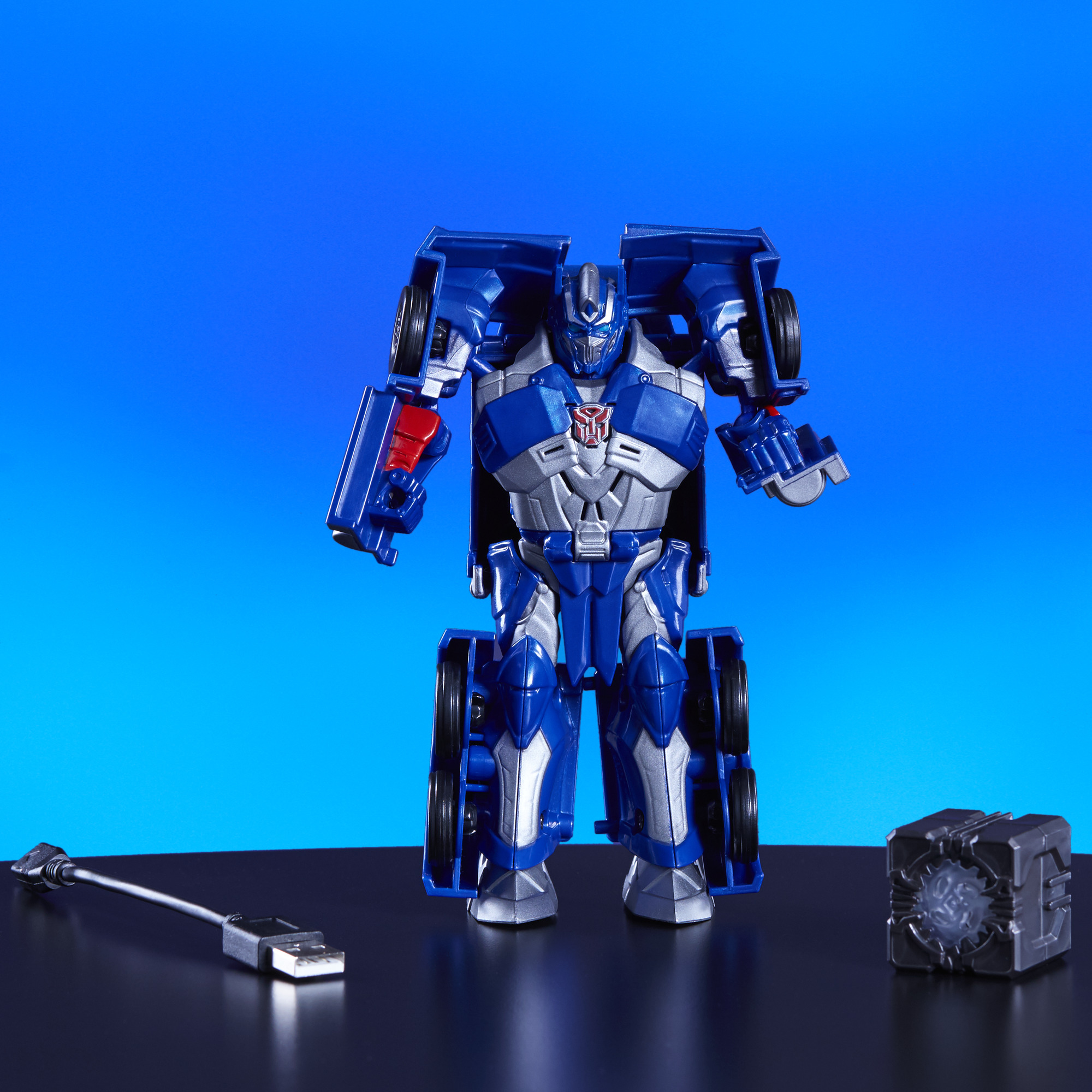 Un robot Transformer blu con accenti argentati, un cavo e una scatola nera su una superficie nera.