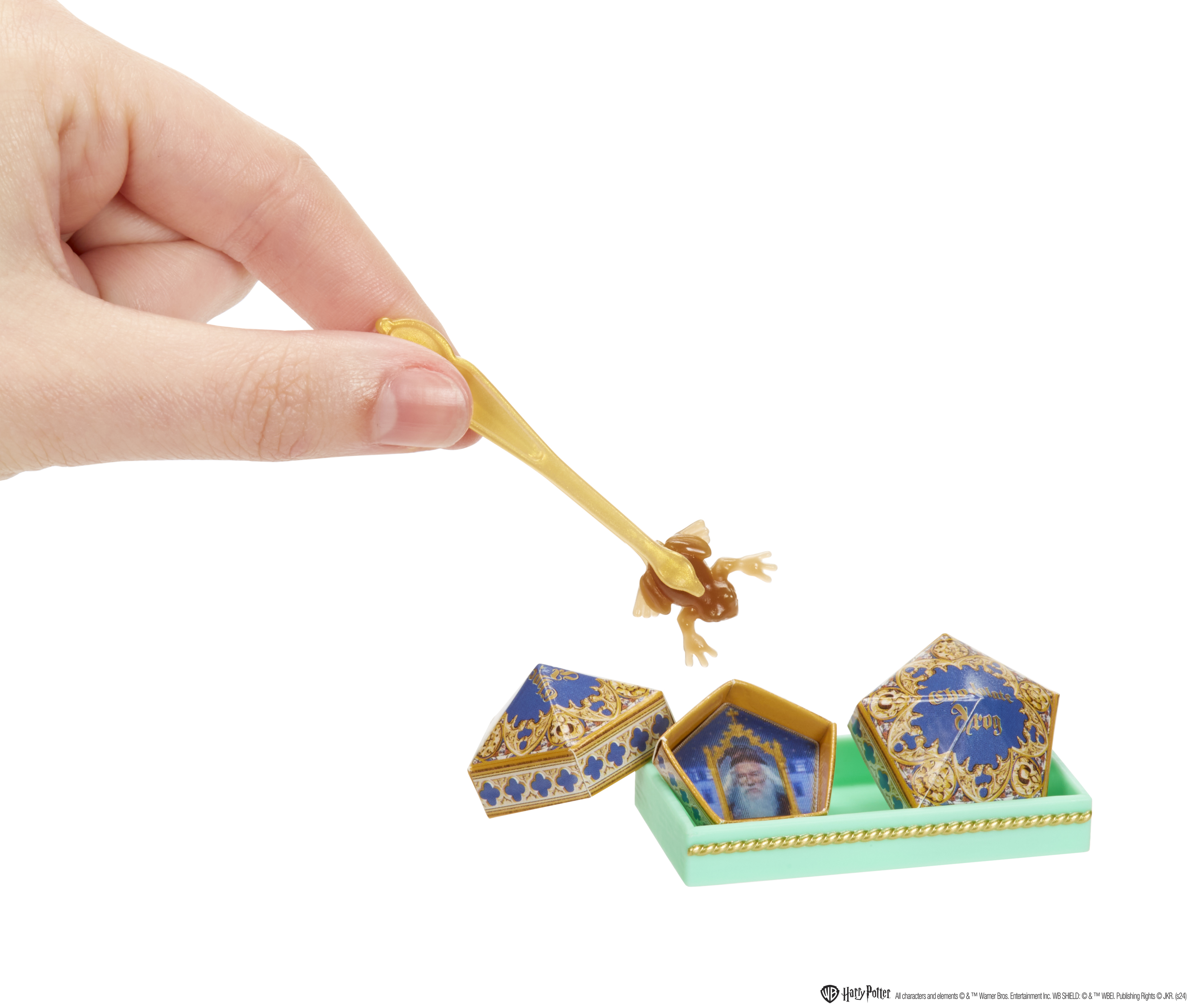 Ręka trzymająca złotą pęsetę, podnosząca żabę z cukierków. W pobliżu znajdują się trzy pudełka z cukierkami.