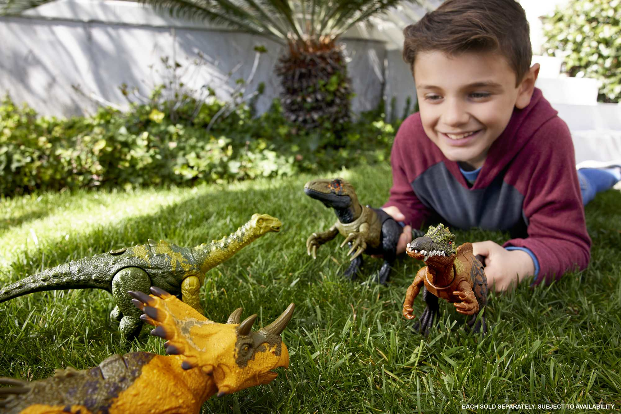 Un ragazzo sorride ai giocattoli di dinosauri sull'erba. Quattro giocattoli di dinosauri sono nella scena.