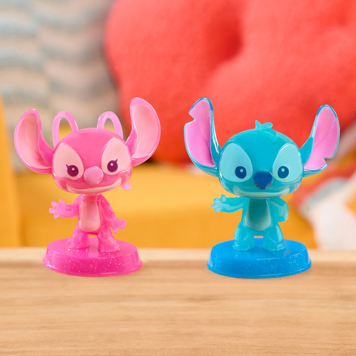 Dwie figurki Stitch, jedna różowa, druga niebieska. Różowa ma różową podstawę, niebieska ma niebieską. Żółte tło.