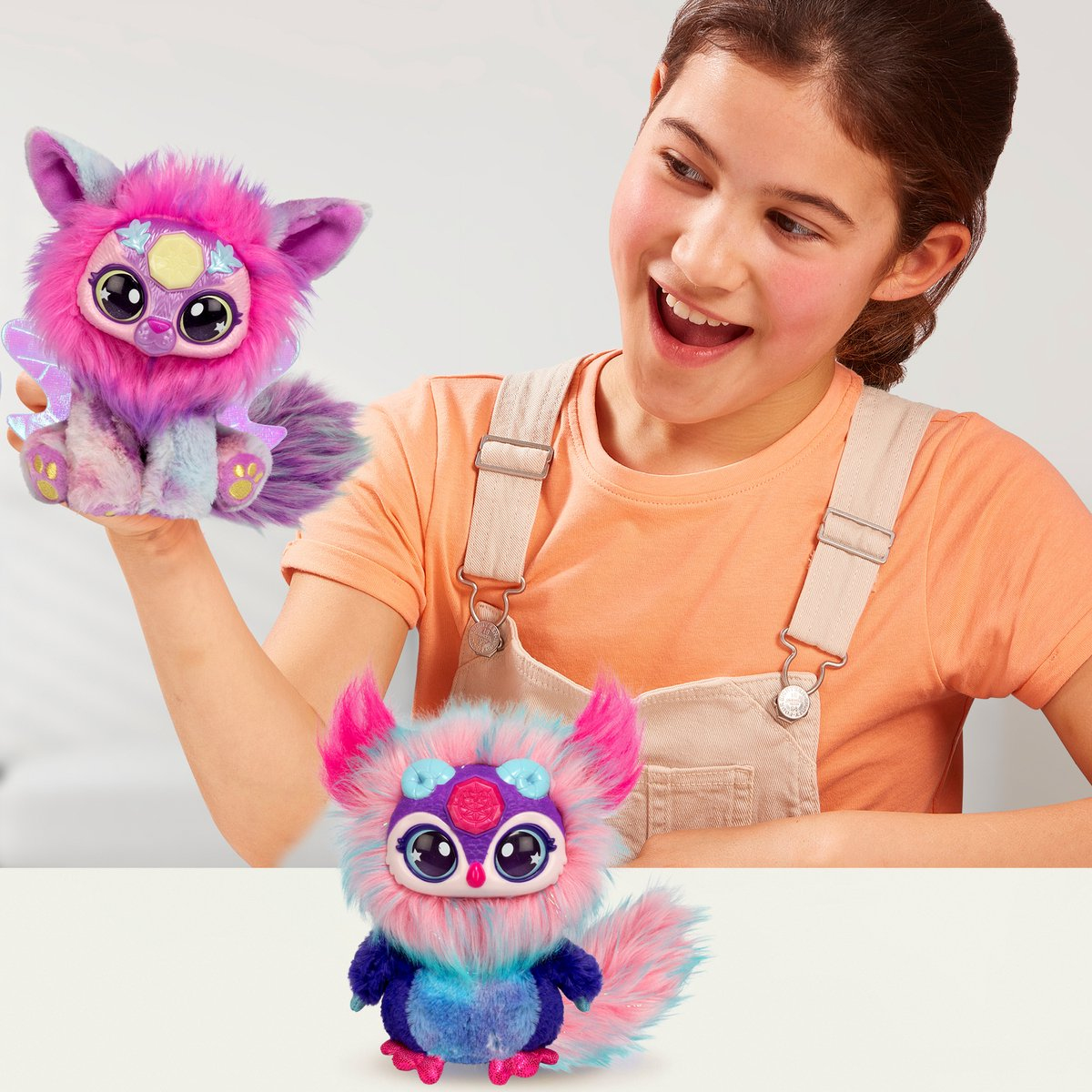 Mädchen lächelt über zwei Plüschtiere: ein rosa-lila Monster und ein blau-rosa Monster auf weißem Untergrund.