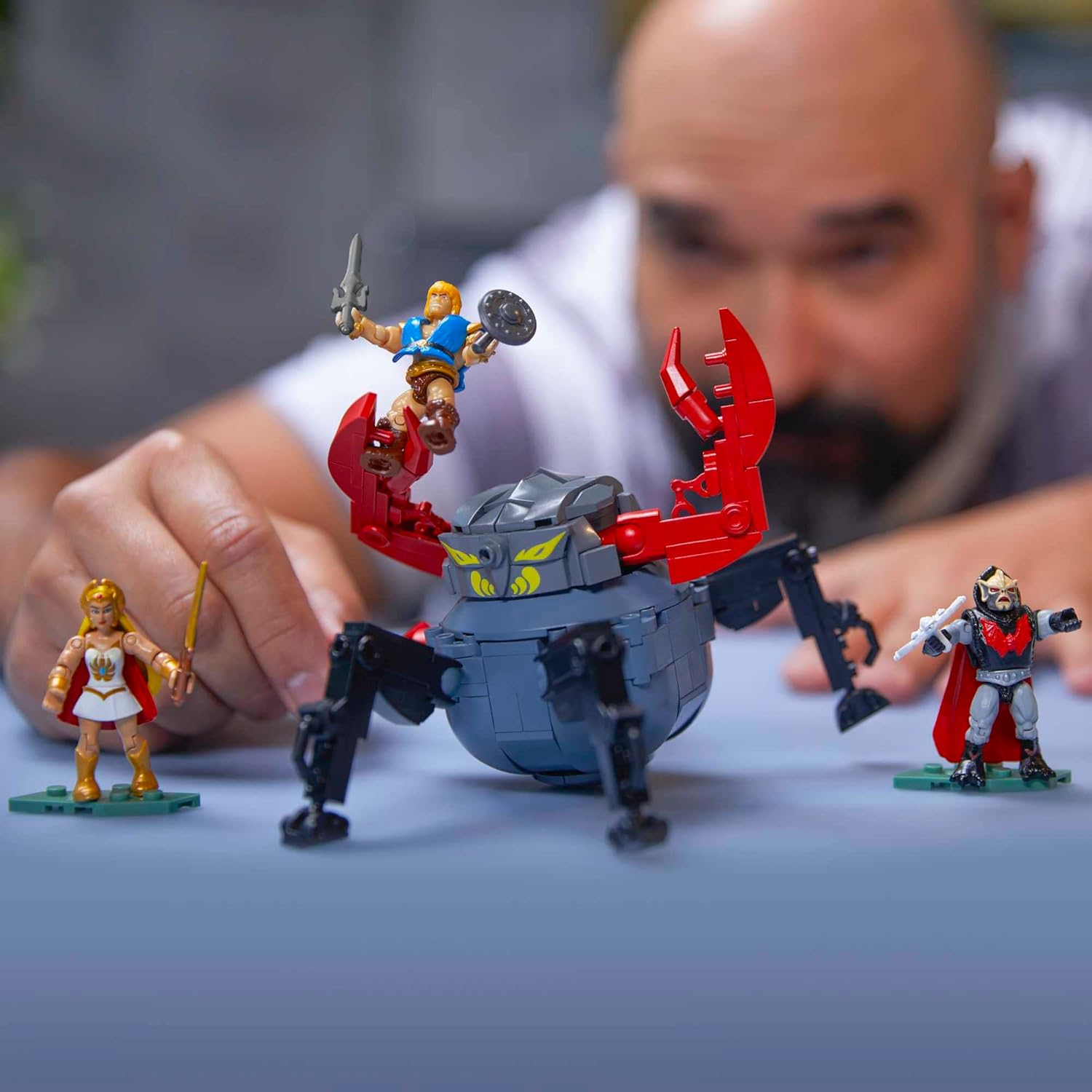 Un hombre juega con figuras de juguete de He-Man, She-Ra y un monstruo parecido a un cangrejo. Están sobre una superficie azul.