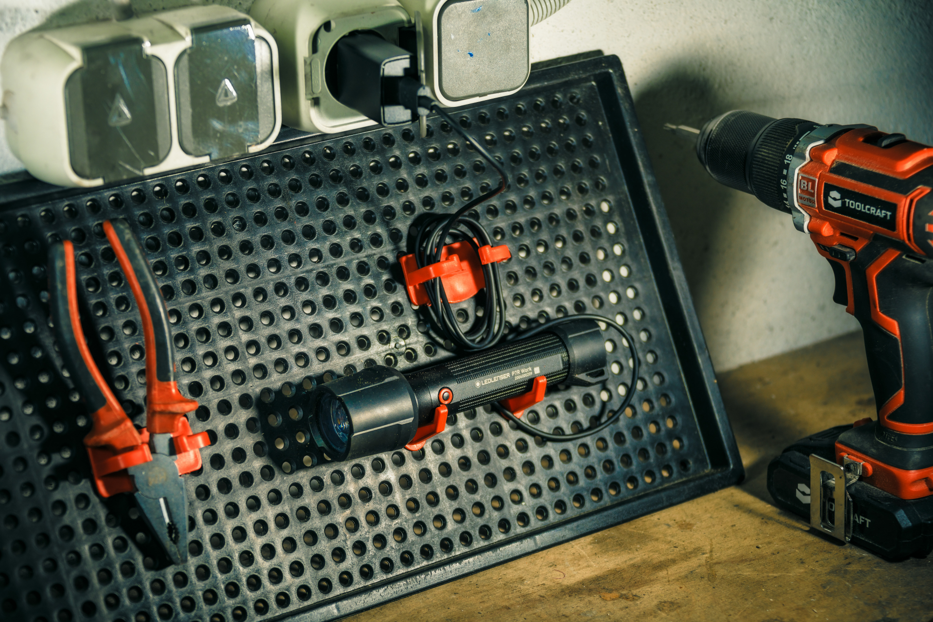 Czarna tablica perforowana z narzędziami: szczypce, latarka, wiertarka. Ustawienie to warsztat z gniazdkami.