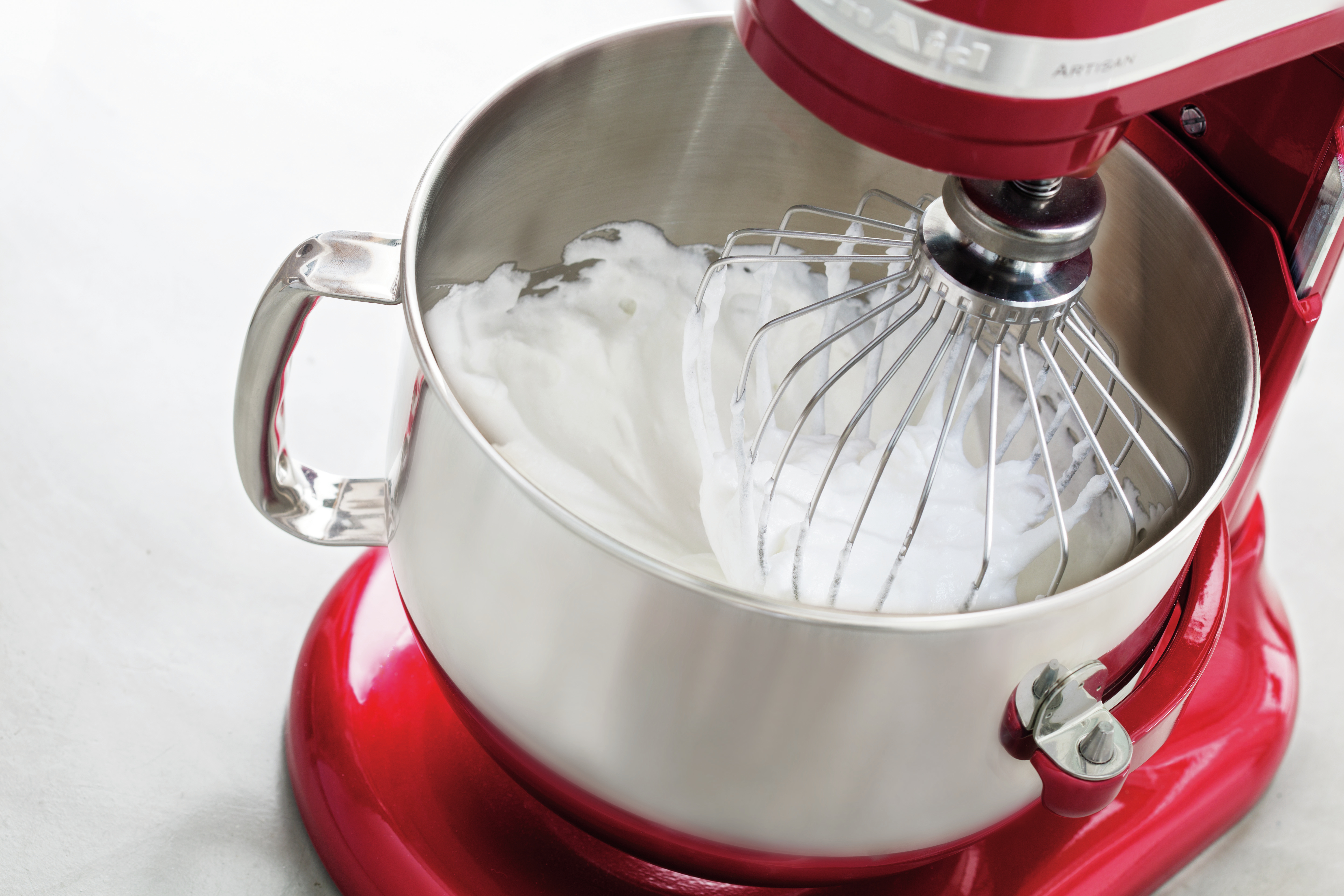 Ein roter KitchenAid-Mixer mit Schneebesenaufsatz, der weiße, fluffige Eier in einer Schüssel verrührt.