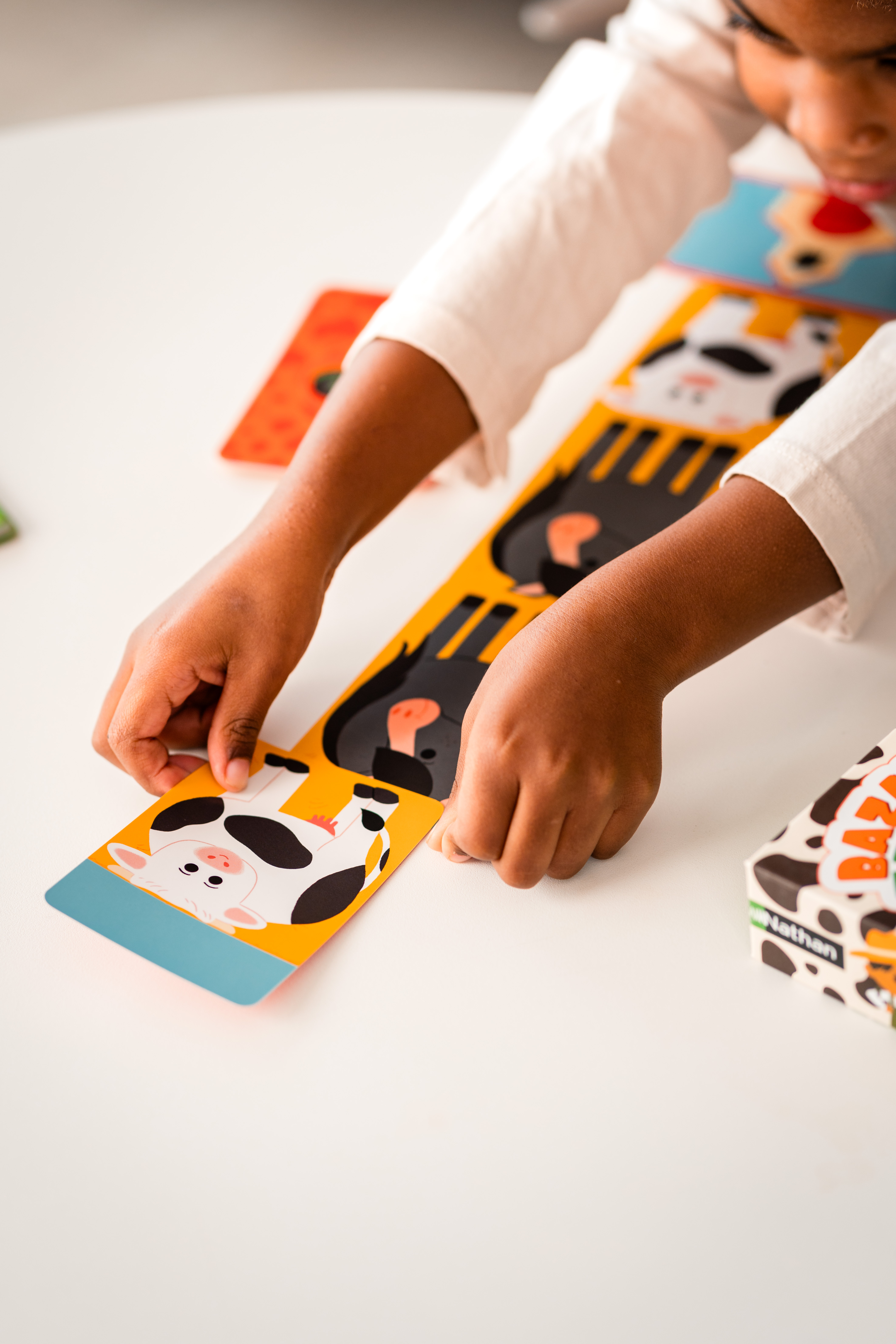 Ręce dziecka składające układankę z krową na białym stole. W pobliżu inne elementy układanki.
