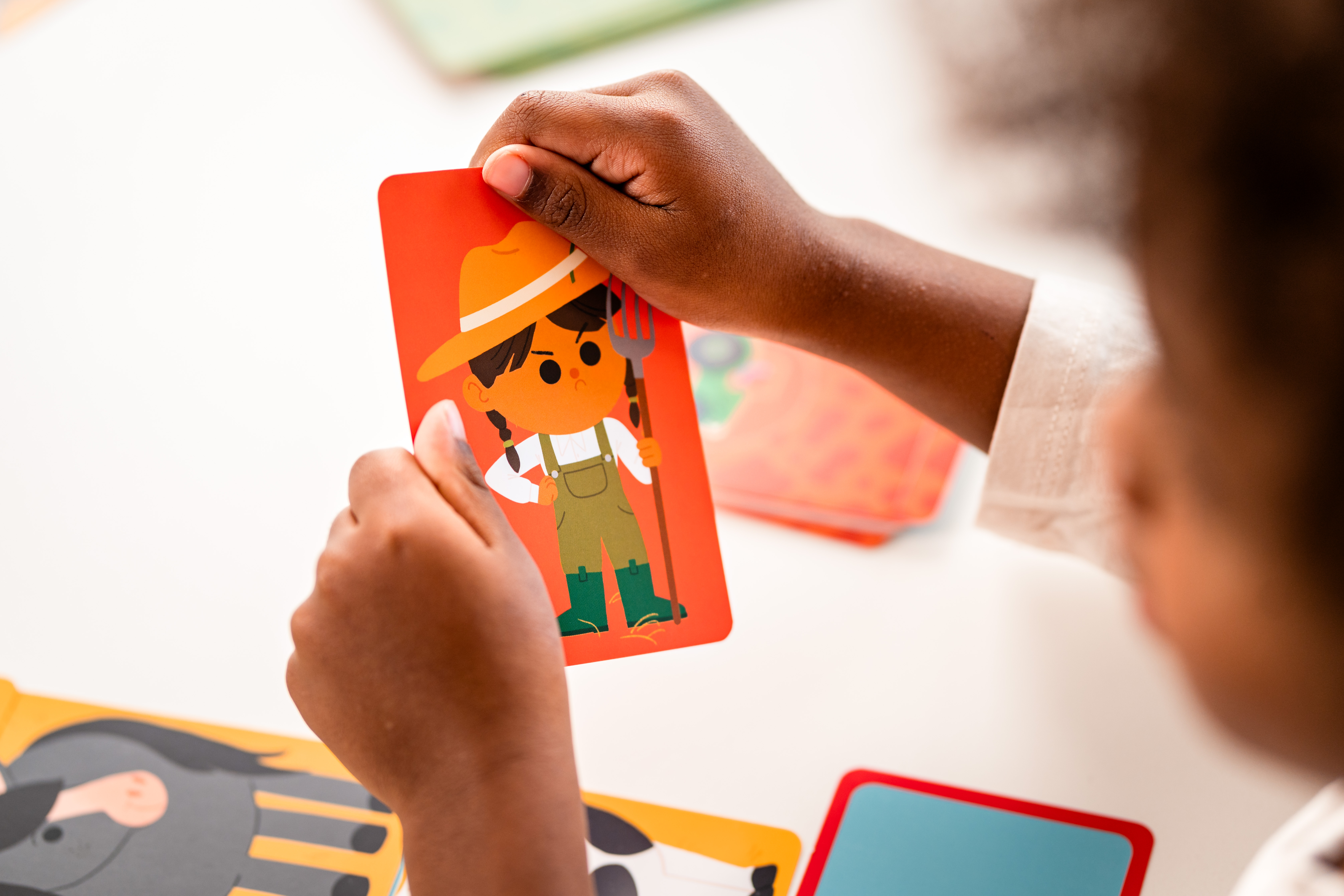 Dziecko trzymające kartę z rolnikiem z kreskówki. Inne karty na białej powierzchni.